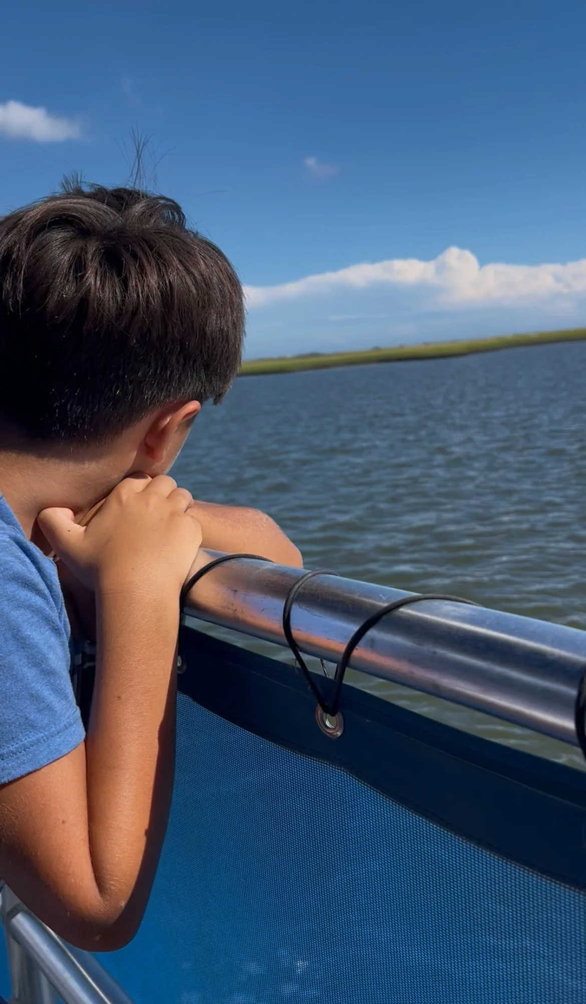 A young boy leaning on a boat railing, looking out over a body of water with a landscape in the distance under a blue sky with clouds.