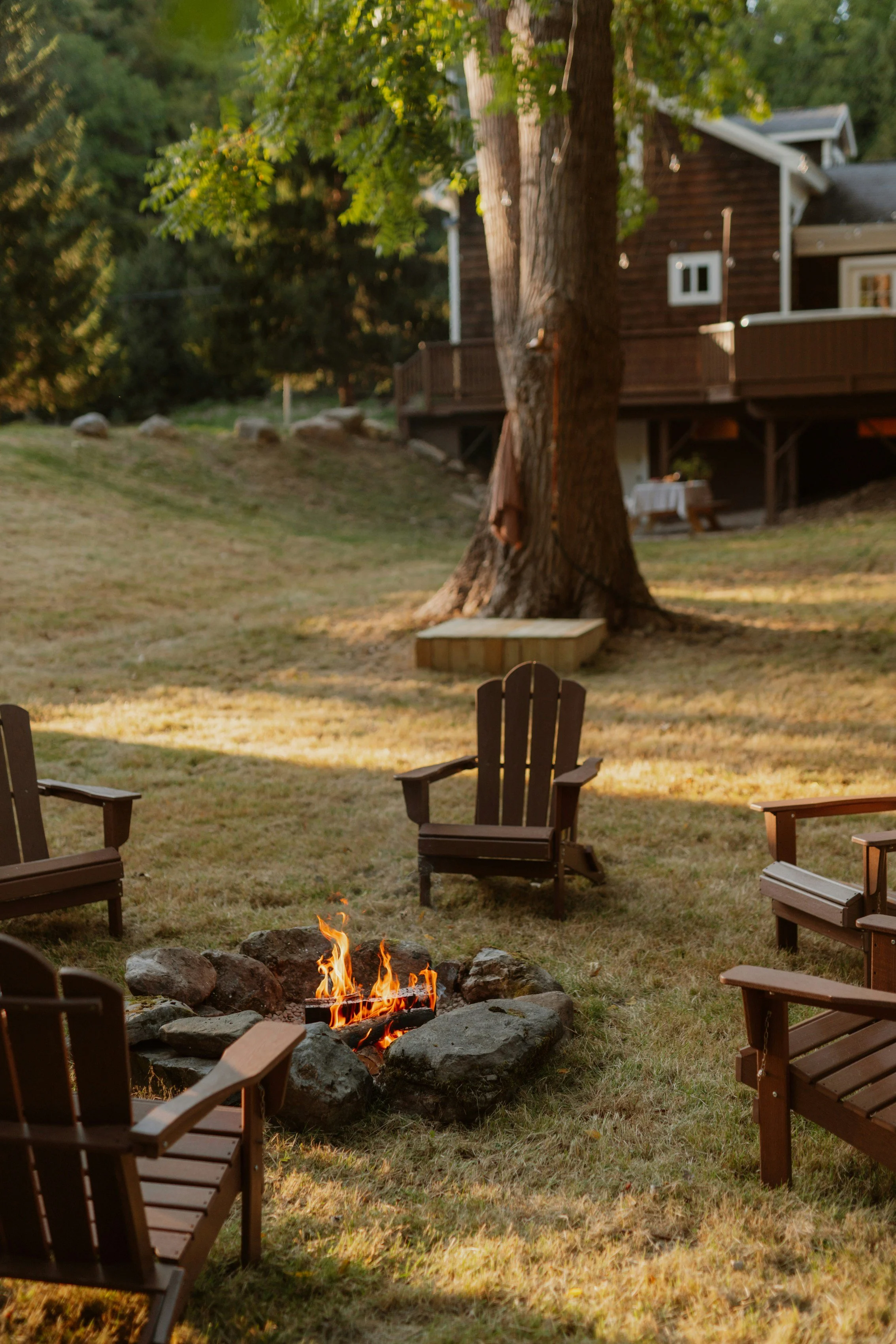 A backyard with a campfire surrounded by five wooden Adirondack chairs, trees, and a house in the background.