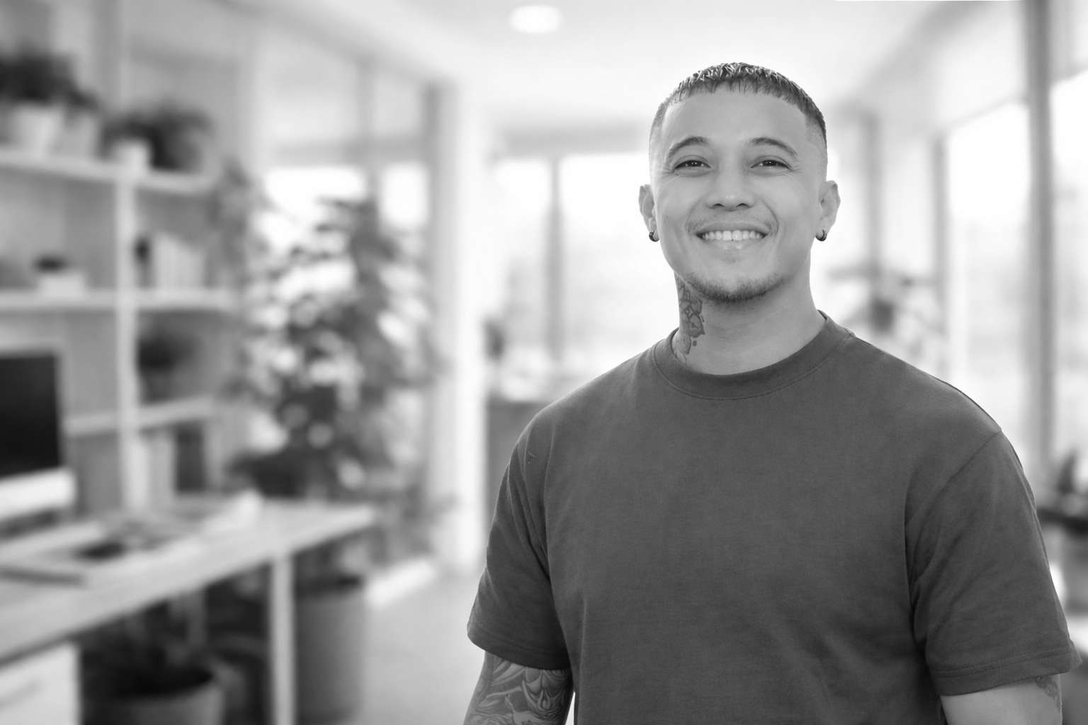 A young man with tattoos smiling in a well-lit room with shelves and a computer in the background.