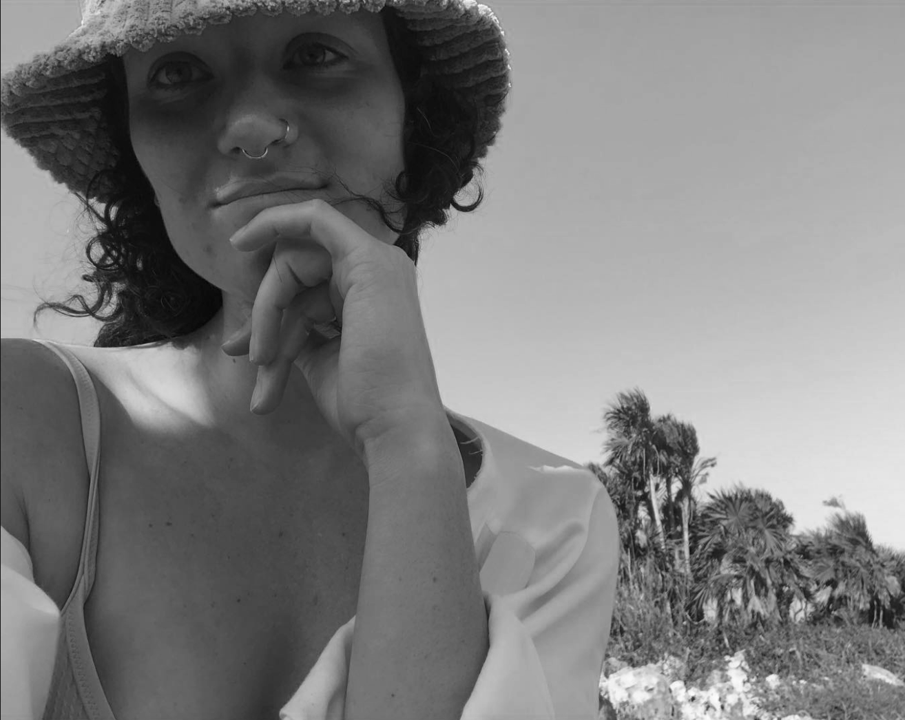 A woman with curly hair, wearing a wide-brimmed hat and a nose ring, touching her chin with her finger. In the background, there are palm trees and a clear sky.