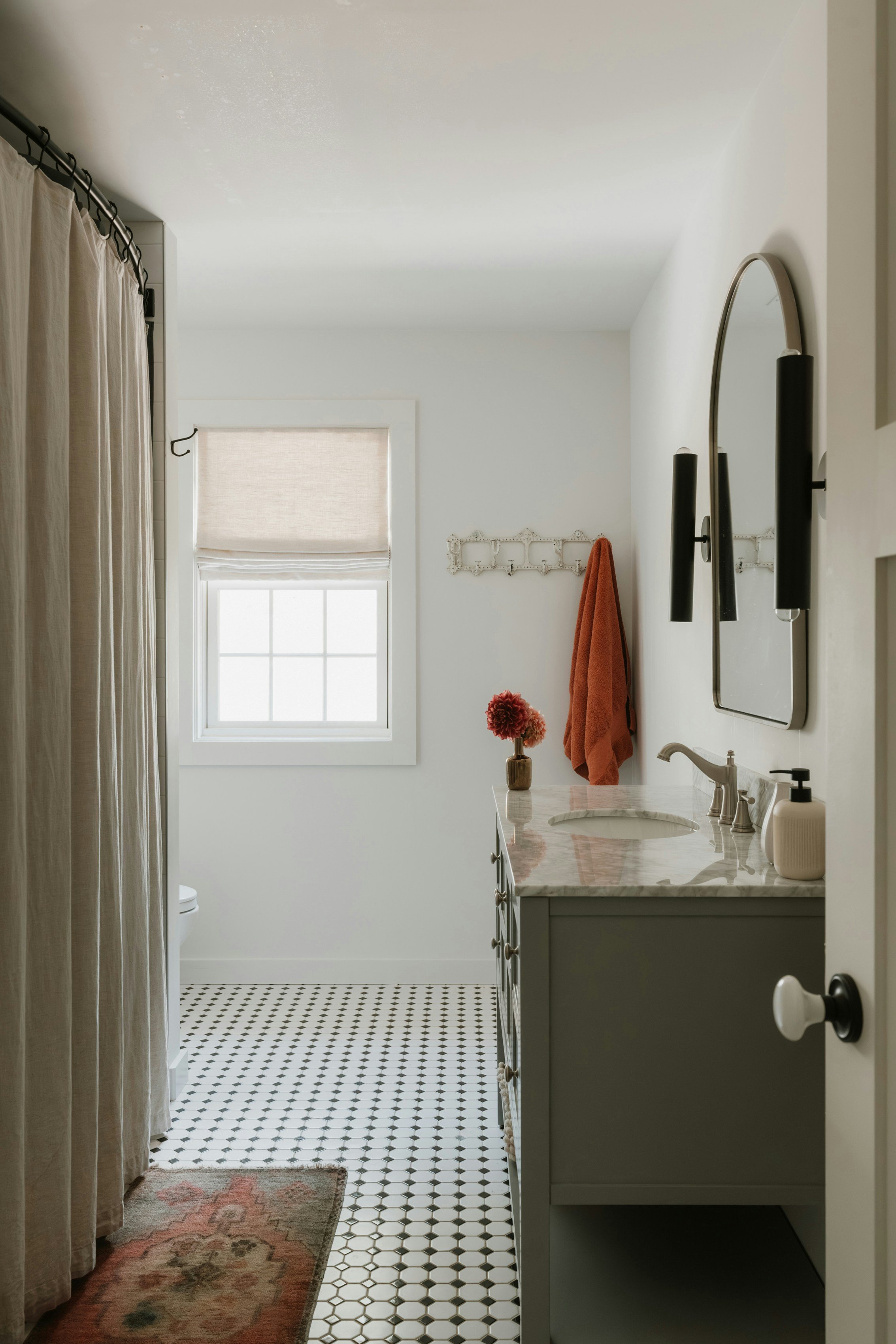 A minimalist bathroom with white walls, a patterned black and white tile floor, a window with a fabric shade, a vanity with a marble countertop, a sink, and a mirror. There is a vase with pink flowers on the counter, a red towel hanging on a wall hook, and a beige shower curtain.