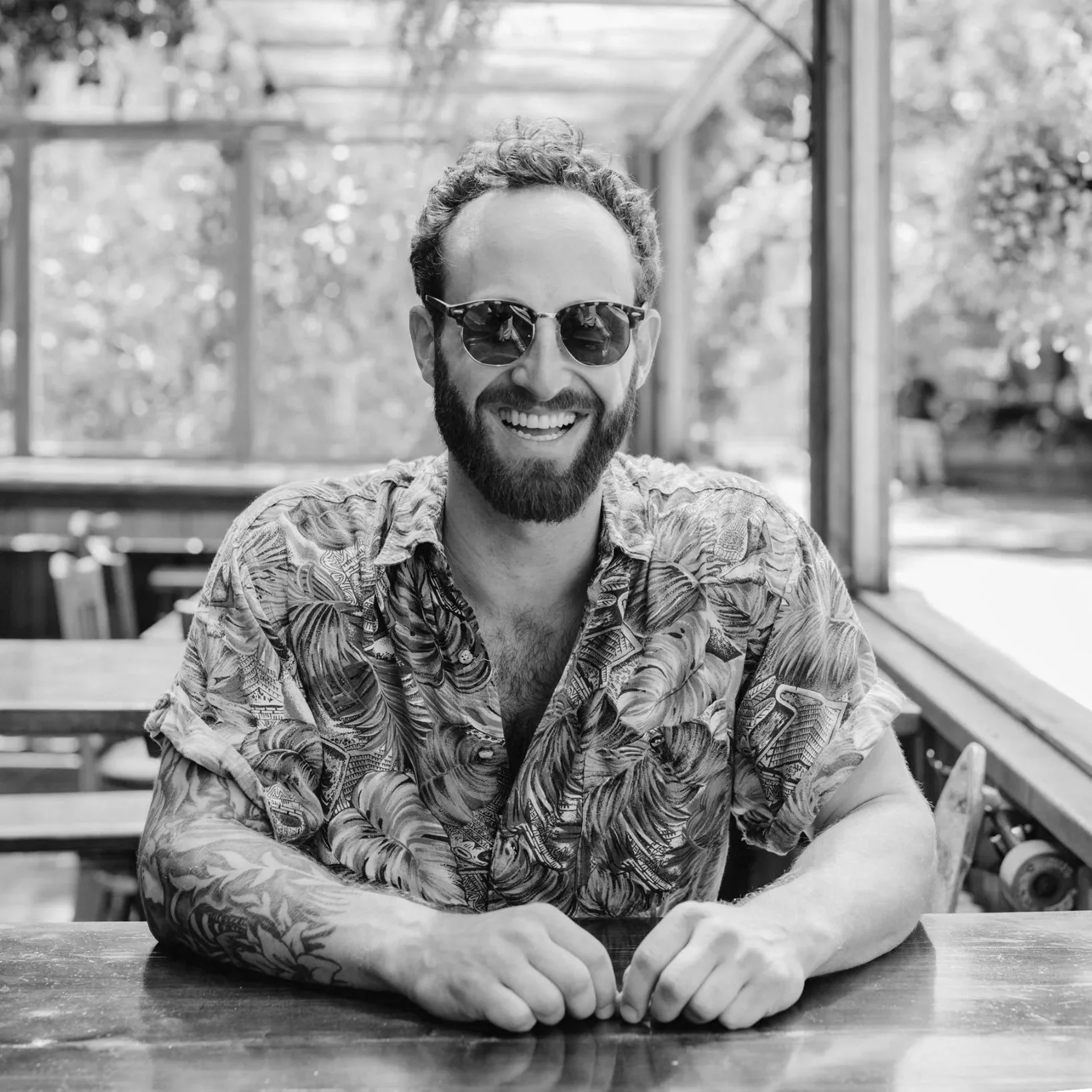A smiling man with a beard and sunglasses sitting at a wooden table in a sunlit outdoor area, wearing a patterned short-sleeve shirt.