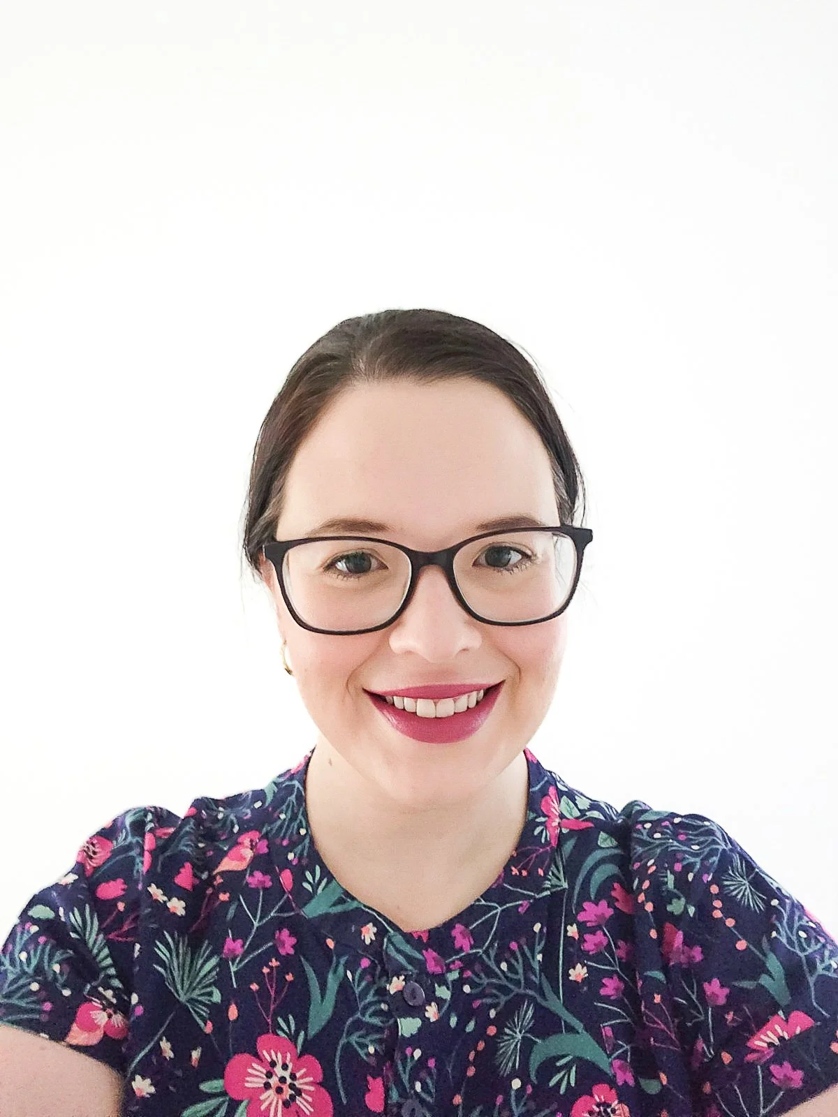 A woman with short dark hair, glasses, and a colorful floral dress smiling against a white background.