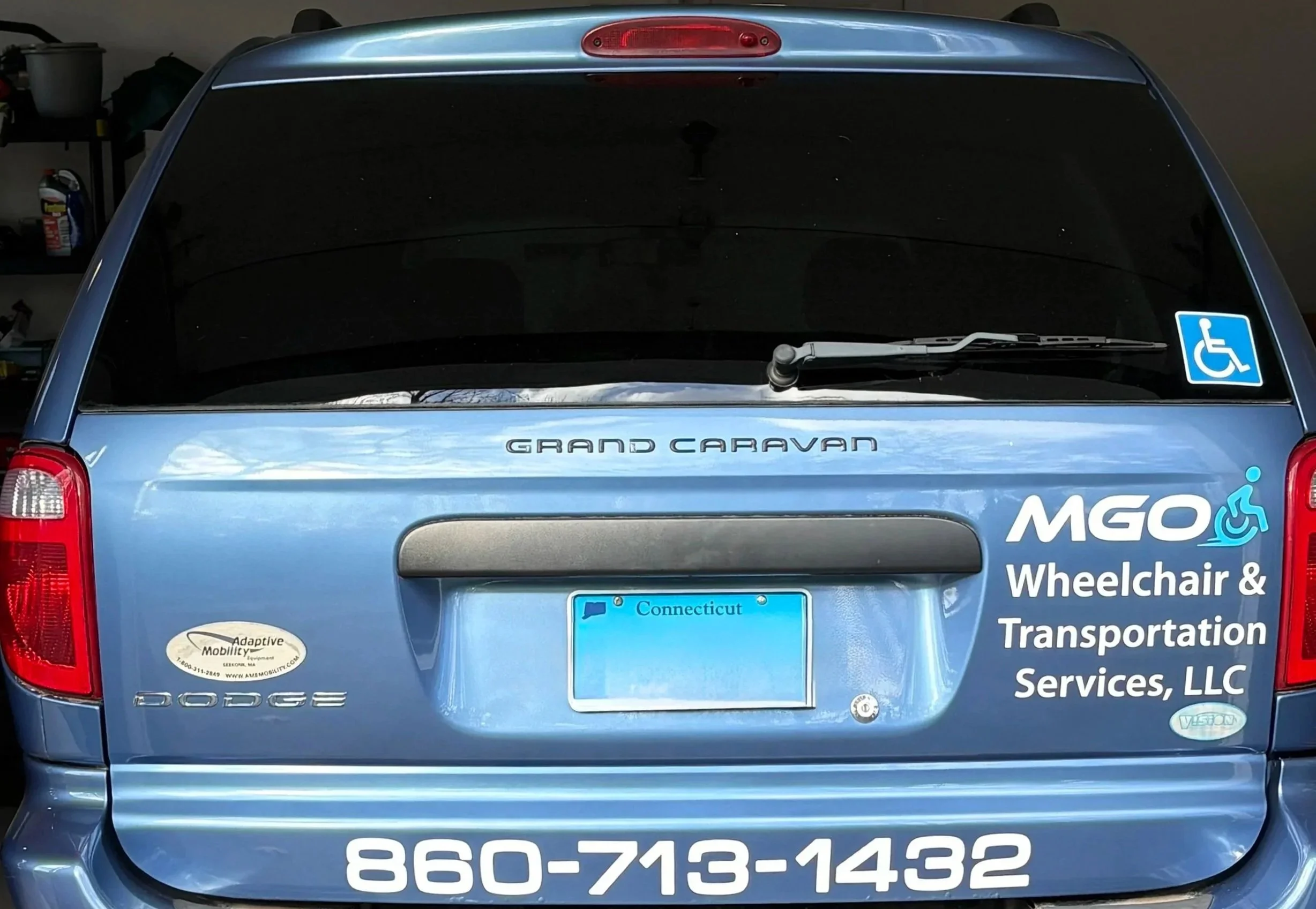 The back of a blue Dodge Grand Caravan minivan has a sticker for MGO Wheelchair & Transportation Services, LLC, with a wheelchair accessible symbol, a Connecticut license plate, and a phone number 860-713-1432.