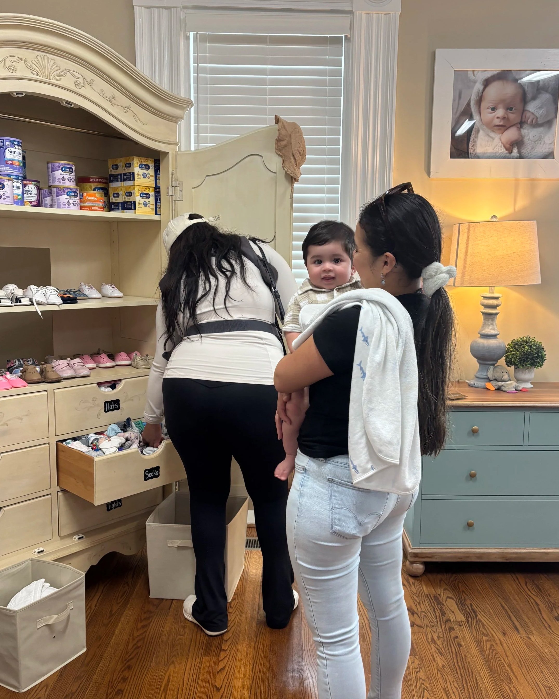 A woman holding a young child at a shoe store, with another woman leaning forward beside them, in front of shoe shelves and a framed baby photo on the wall.