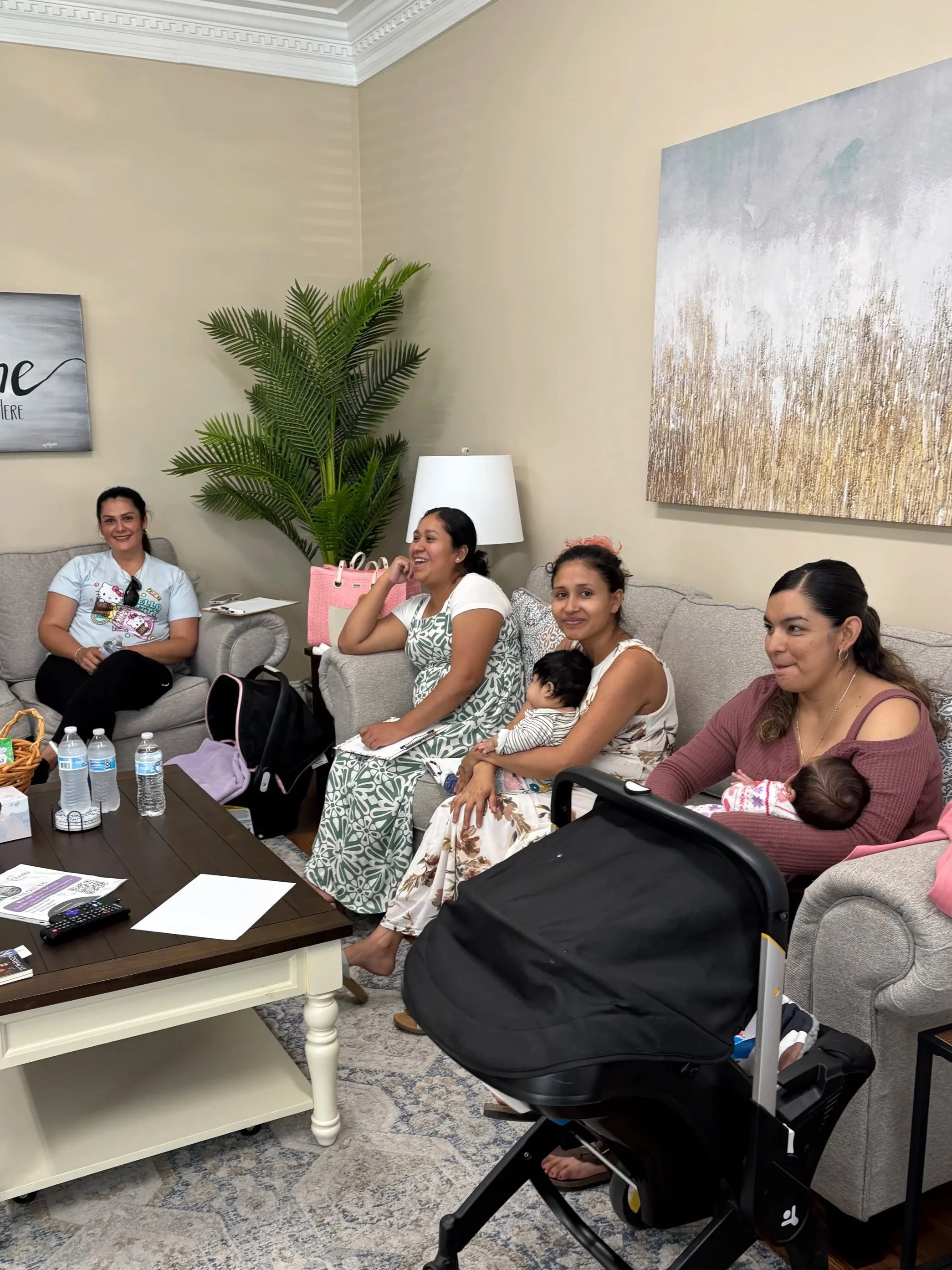 A group of women and children sitting on sofas in a living room, smiling and engaged in conversation. There is a large potted plant, a table with water bottles, and wall art in the background.