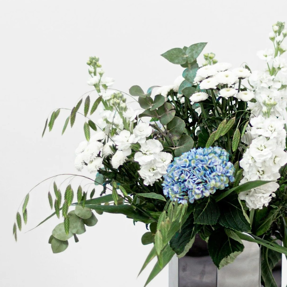 A floral arrangement in a black vase, featuring white flowers, blue hydrangeas, and green foliage.