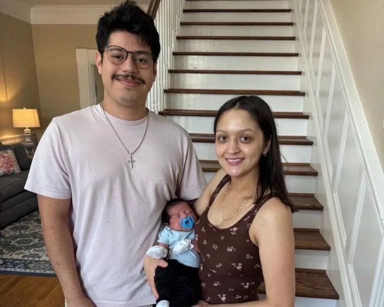 A smiling couple holding a newborn baby in front of a staircase inside a home. The man has dark hair, glasses, and a mustache, wearing a light-colored t-shirt with a necklace. The woman has long dark hair, wearing a brown sleeveless top. The baby has a pacifier and is dressed in light blue and black.