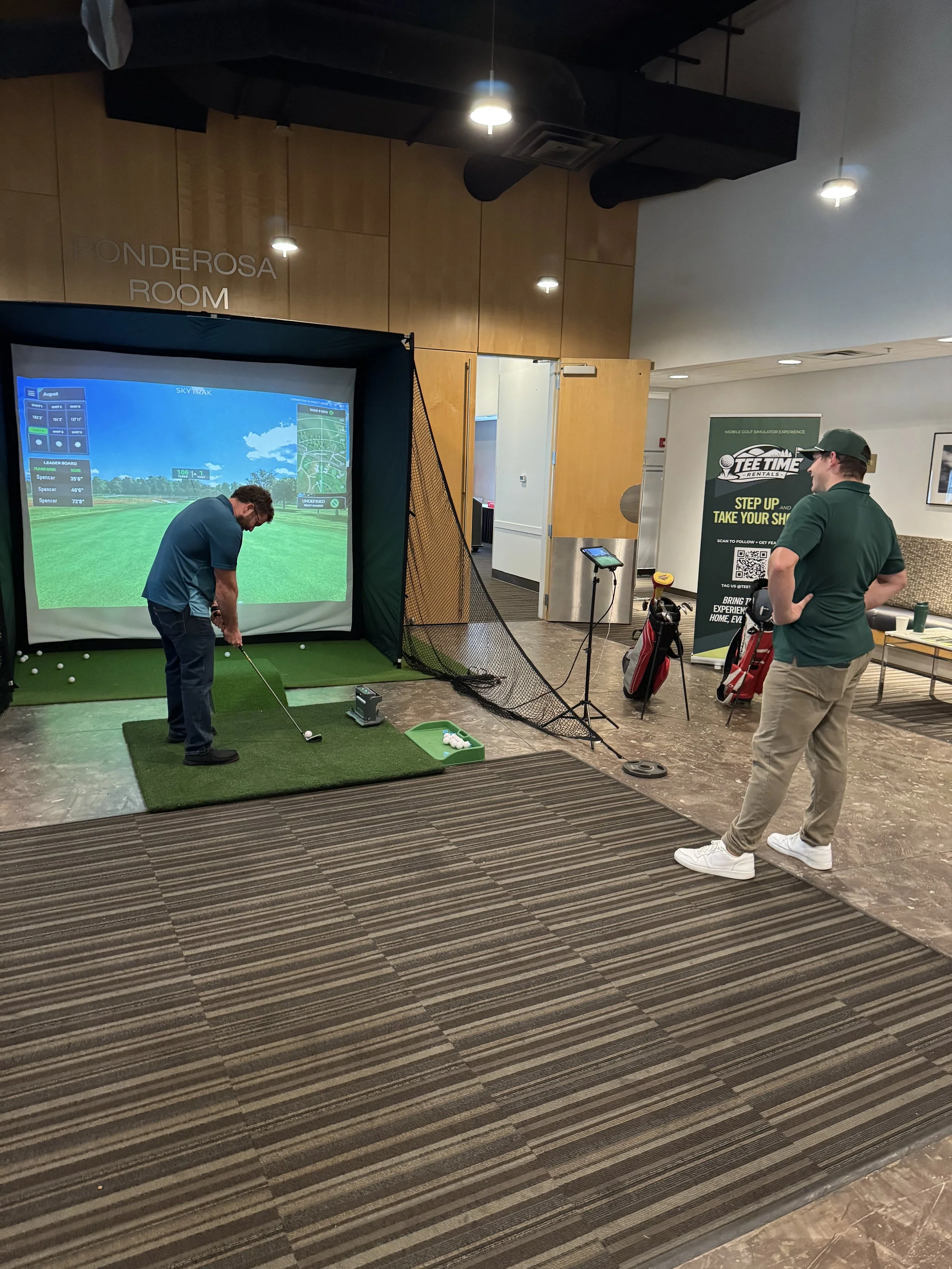 Indoor golf simulator with two men, one swinging a golf club, and another watching. Large screen displays a virtual golf course, and there are golf bags and equipment nearby.