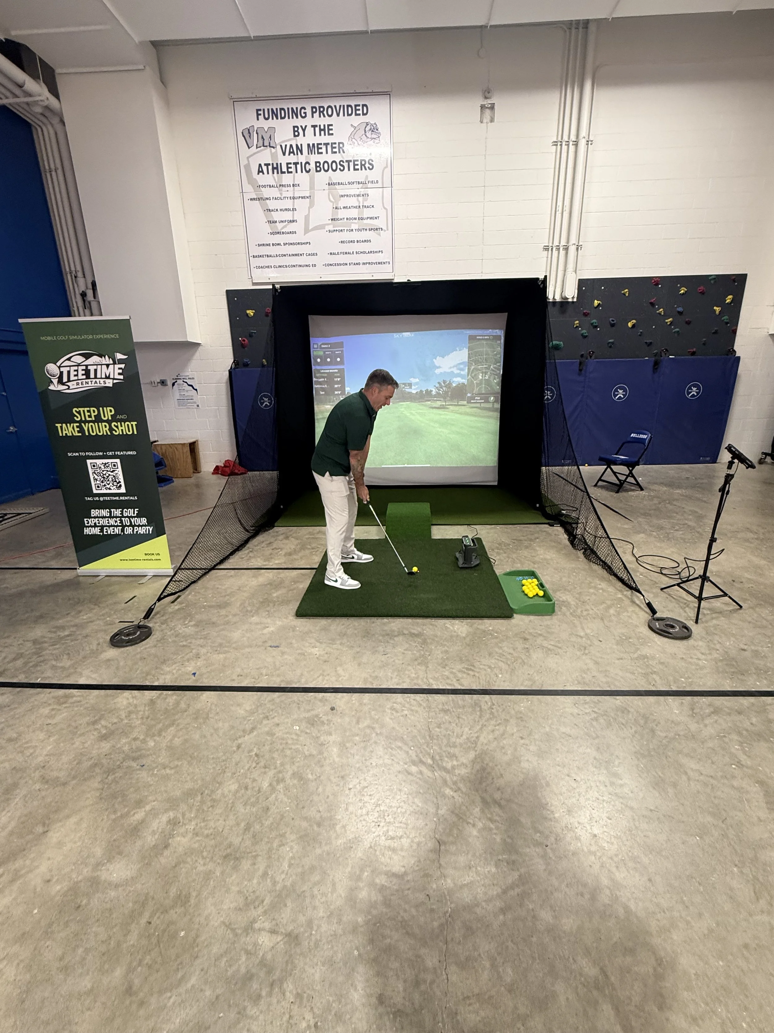 Indoor golf simulation setup with a man preparing to hit a golf ball on a simulated golf course, surrounded by playing mats, a screen displaying a golf course, and promotional banners.
