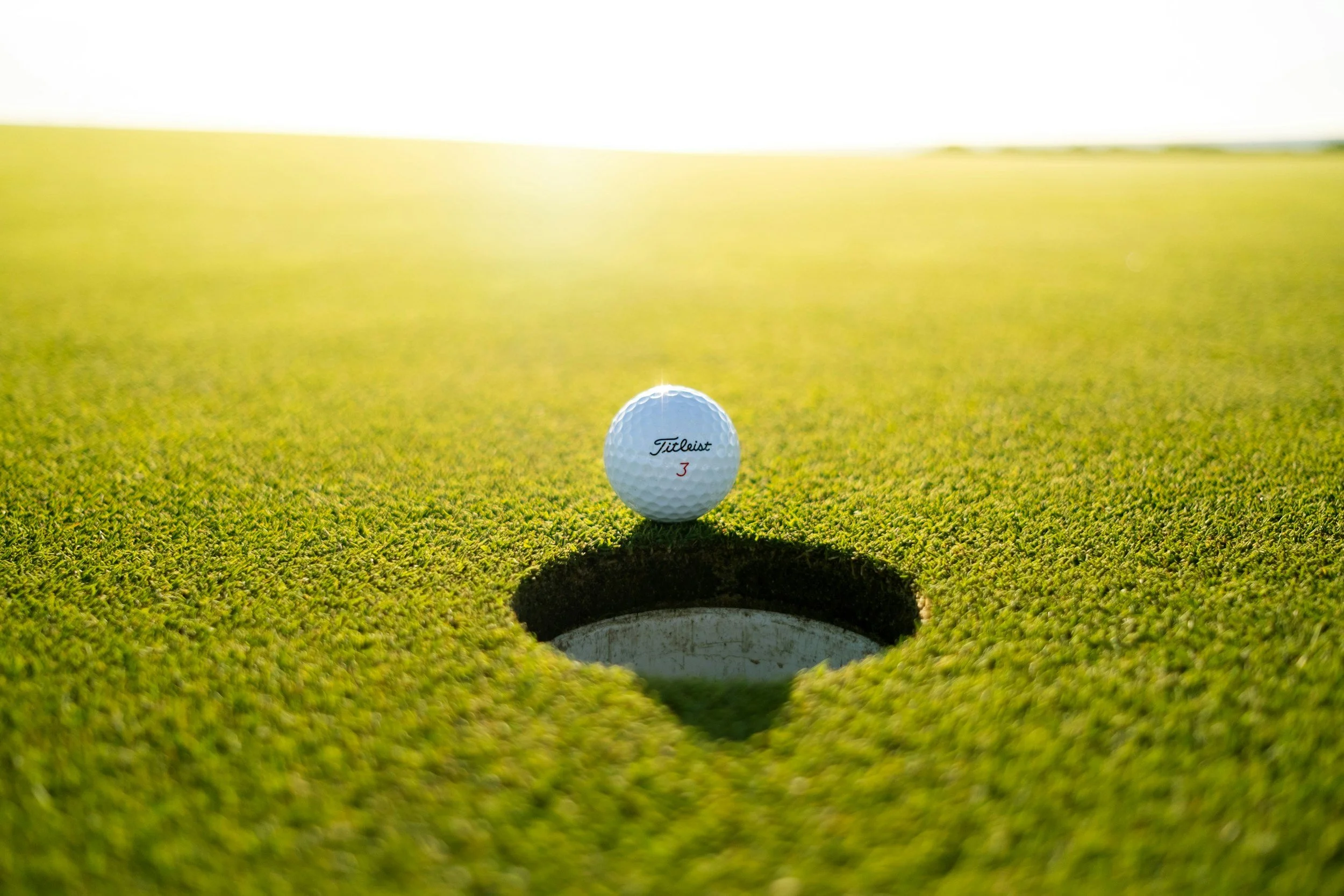 Golf ball positioned near the hole on a putting green, with bright sunlight and a blurred background.