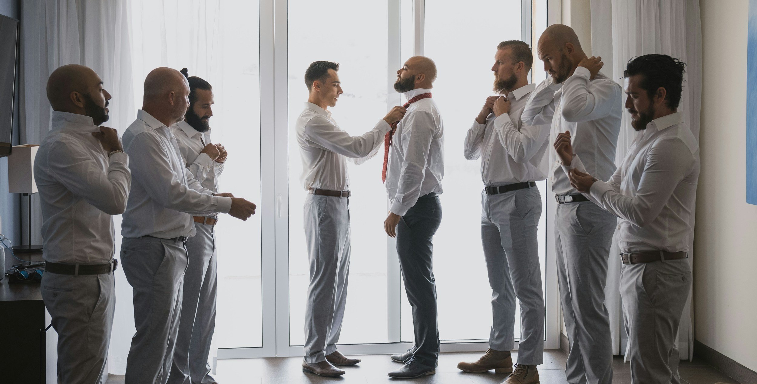 A group of men in white shirts and gray pants standing in a line in front of a window while one man adjusts another man's red tie. The men have different skin tones and beards, and some are adjusting their collars.