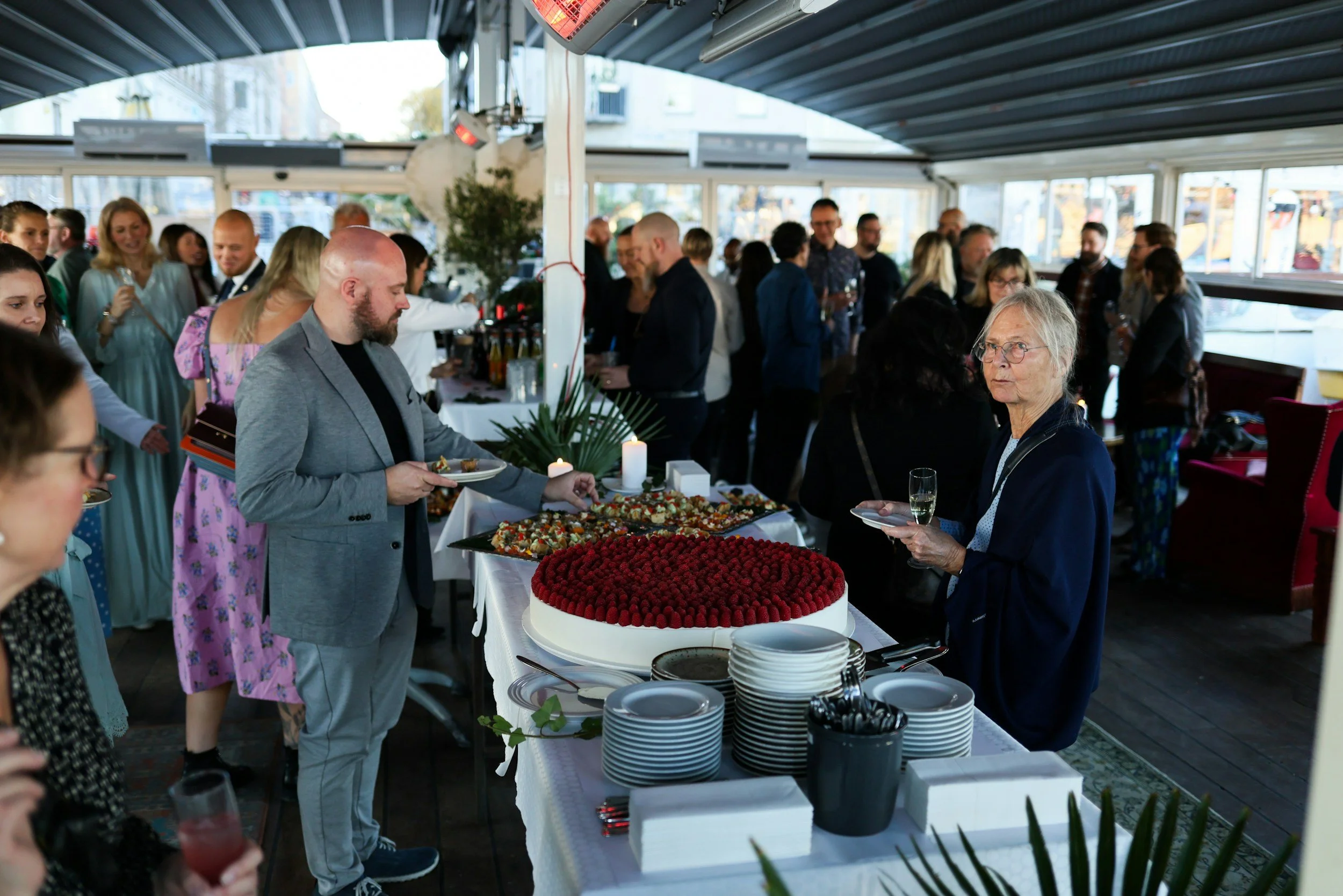 People at a social gathering serving themselves food from a buffet table, there are desserts including a cake topped with raspberries and assorted pastries, in a bright indoor space with large windows.