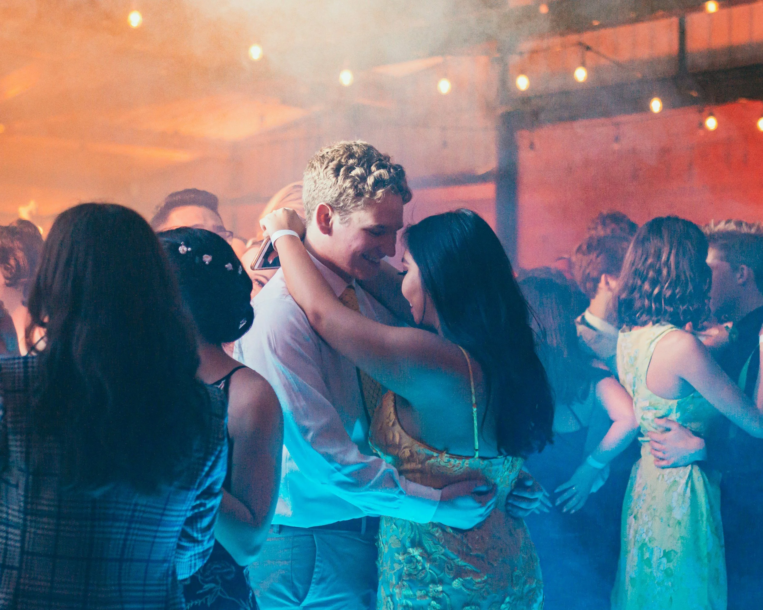 Couple dancing closely at a party with warm lighting and other people in the background.