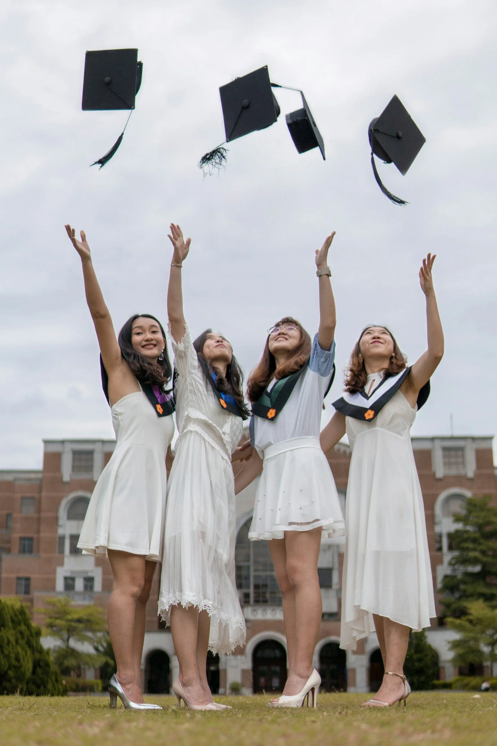 Four women in white dresses and high heels stand together outdoors, celebrating graduation. Their graduation caps are soaring through the air against an overcast sky, and they are smiling with arms raised, in front of a large brick building with arched windows.