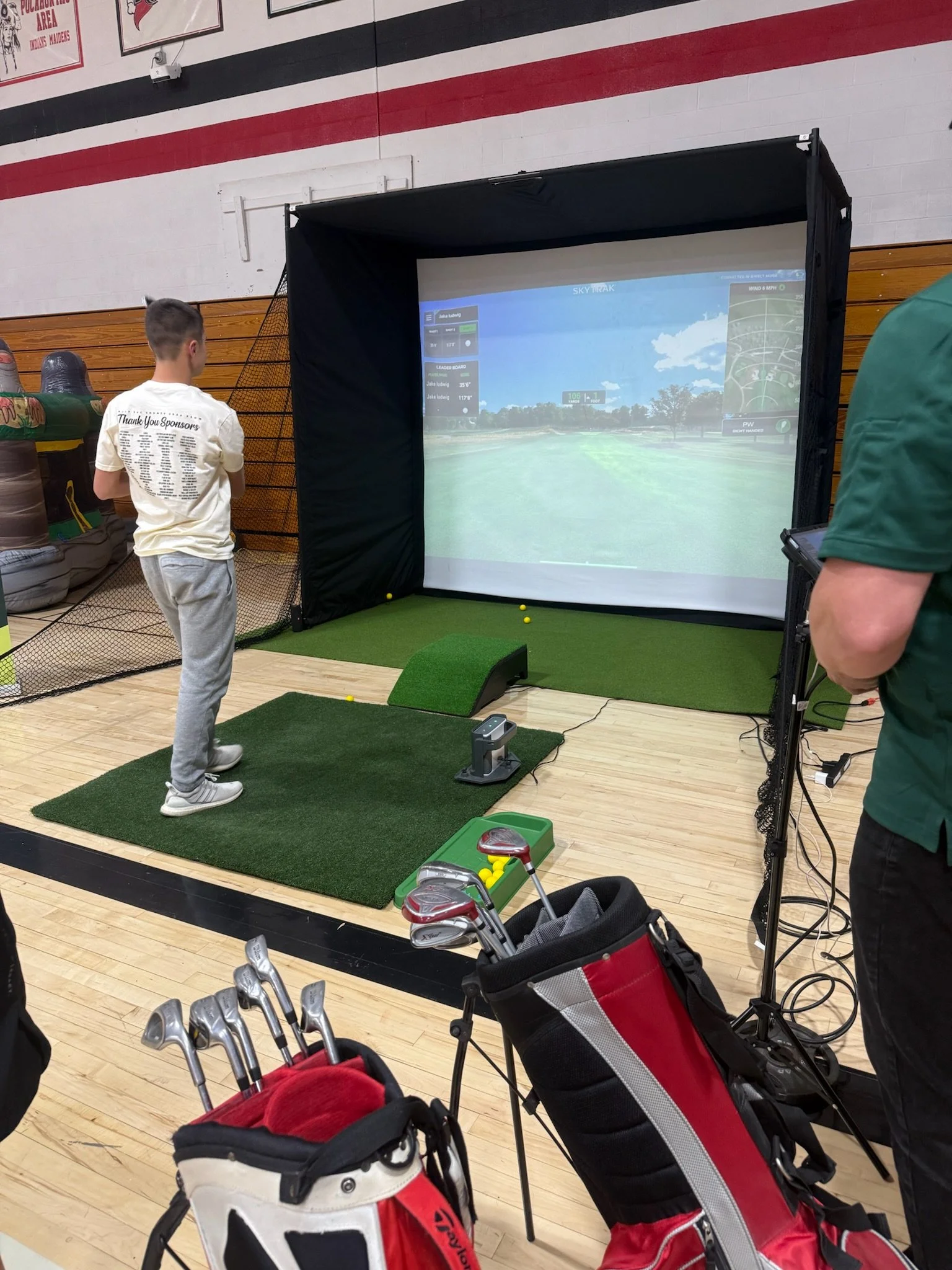 Indoor golf simulation setup with a large screen displaying a virtual golf course, a young boy playing, and golf clubs in golf bags in the foreground.