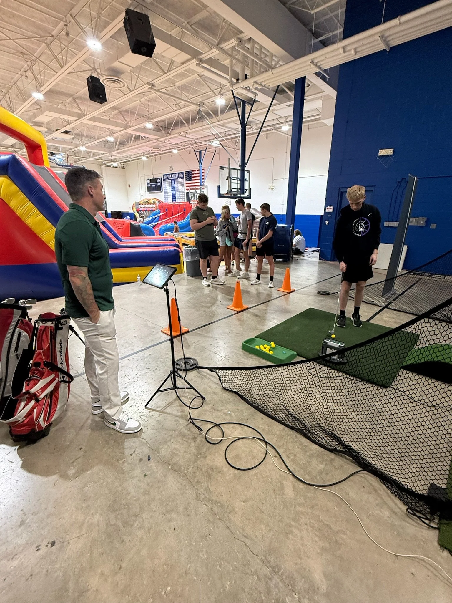 Indoor sports facility with people gathered around a game station. There is an inflatable obstacle course in the background and a golf practice area with yellow balls in the foreground. A person is standing to the left with a golf club, and others are standing in line or preparing to play.