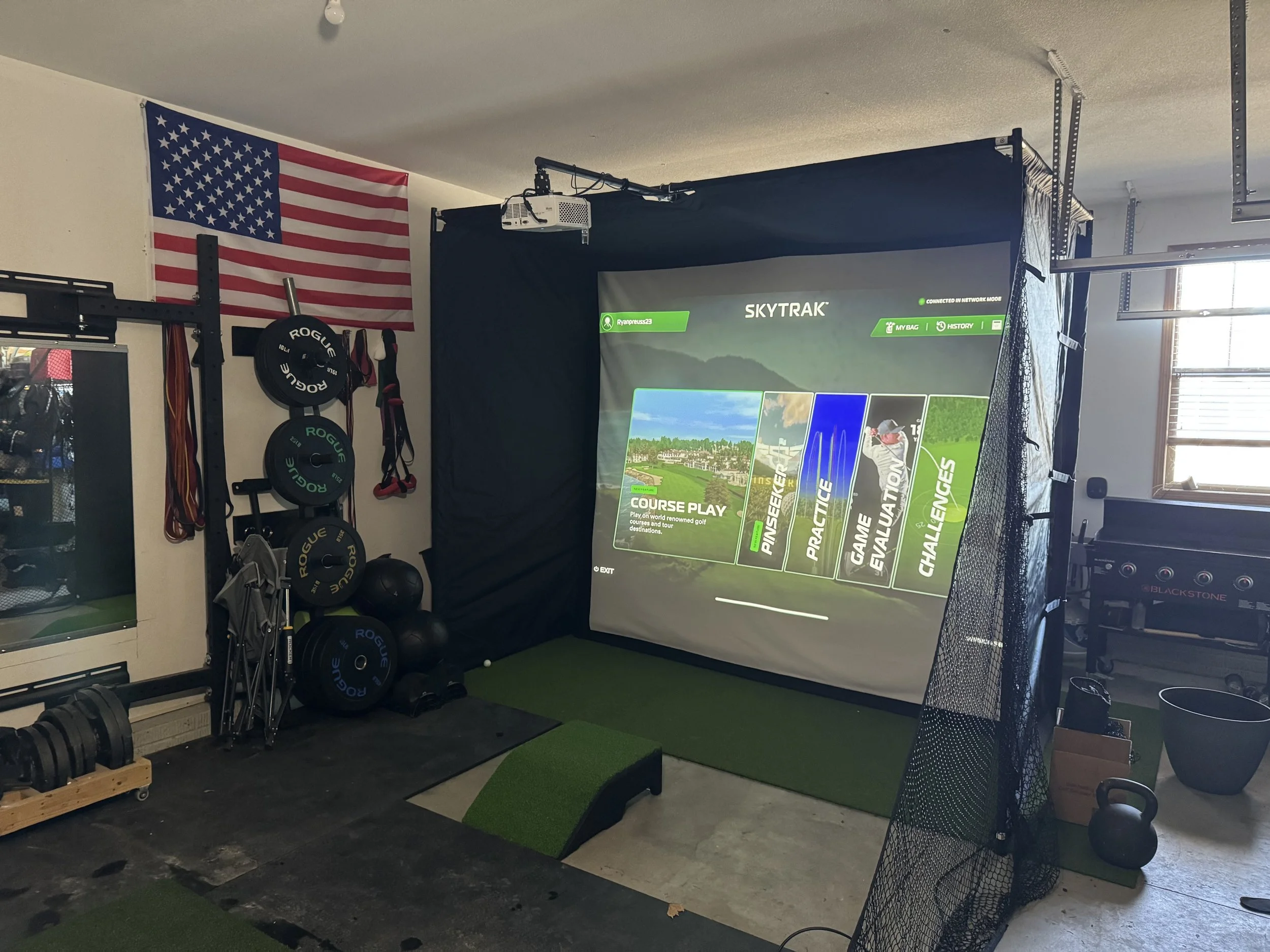 Indoor golf simulator setup with a large screen displaying game options, accompanied by golf equipment including weights, kettlebells, and resistance bands. An American flag hangs on the wall to the left of the setup.