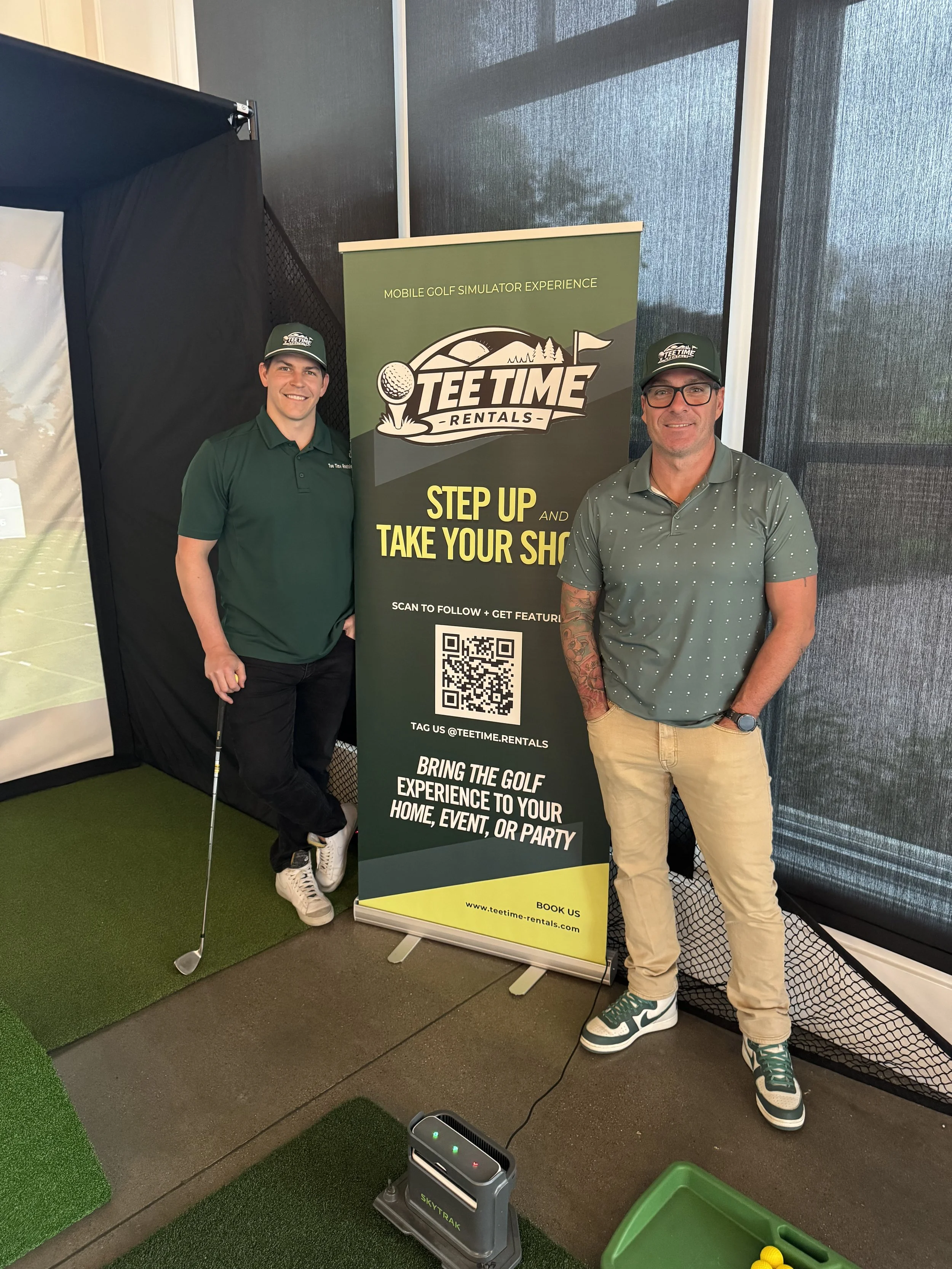 Two men standing beside a vertical banner for Teet Time Rentals in an indoor golf simulation facility. The man on the left is holding a golf club, wearing a green polo shirt, black pants, and a green cap. The man on the right is wearing glasses, a gray dotted polo shirt, beige pants, and teal sneakers. The banner encourages customers to step up and take their shot, with a QR code and website URL at the bottom.