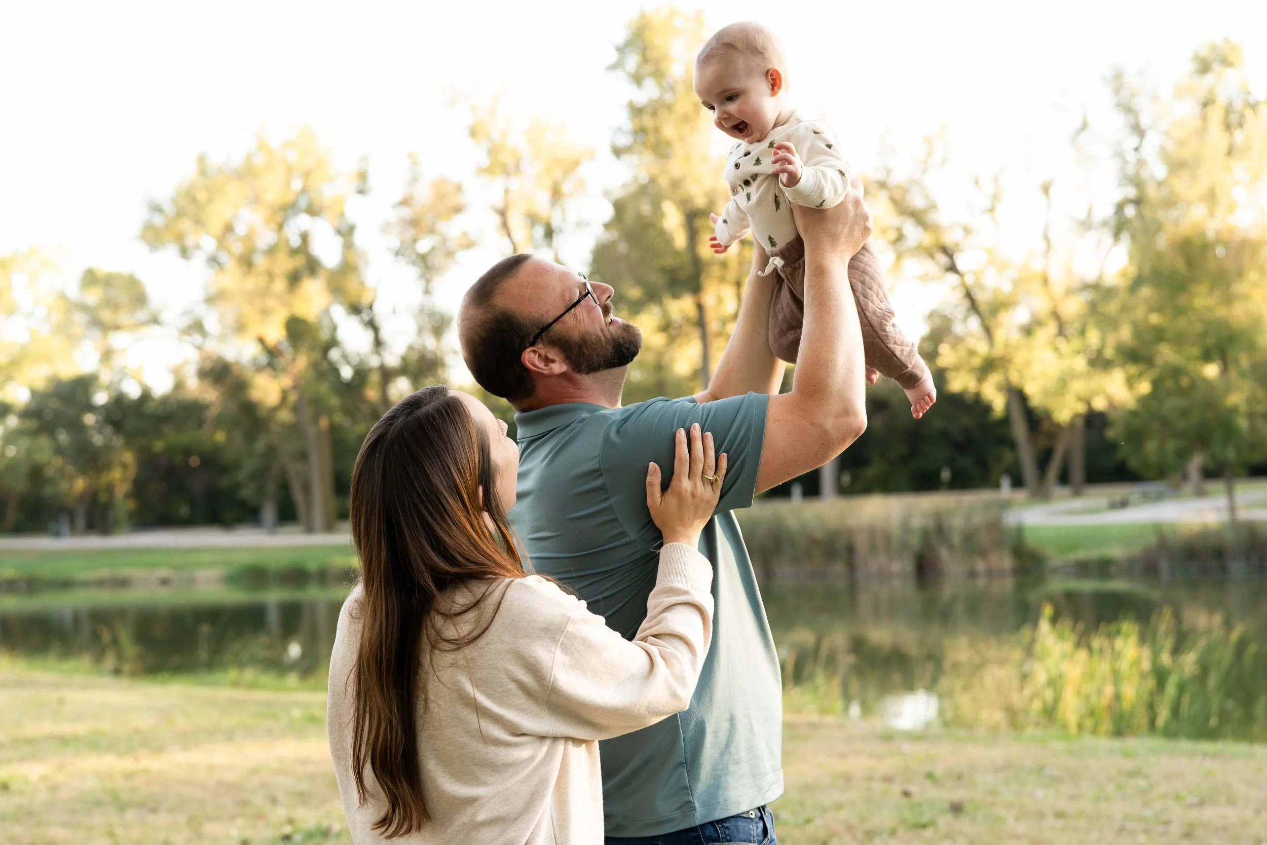 Family outdoors, father lifting a baby girl, mother supports father, trees and lake in background.