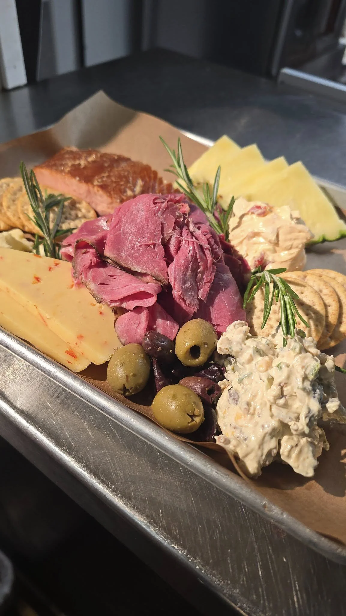 A tray of assorted deli meats, cheeses, olives, and spreads garnished with fresh rosemary sprigs.