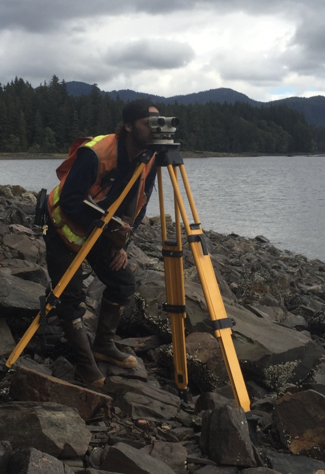 A person in outdoor clothing with orange safety vest looking through a theodolite on a rocky shoreline near a lake, with forested hills and cloudy sky in the background.