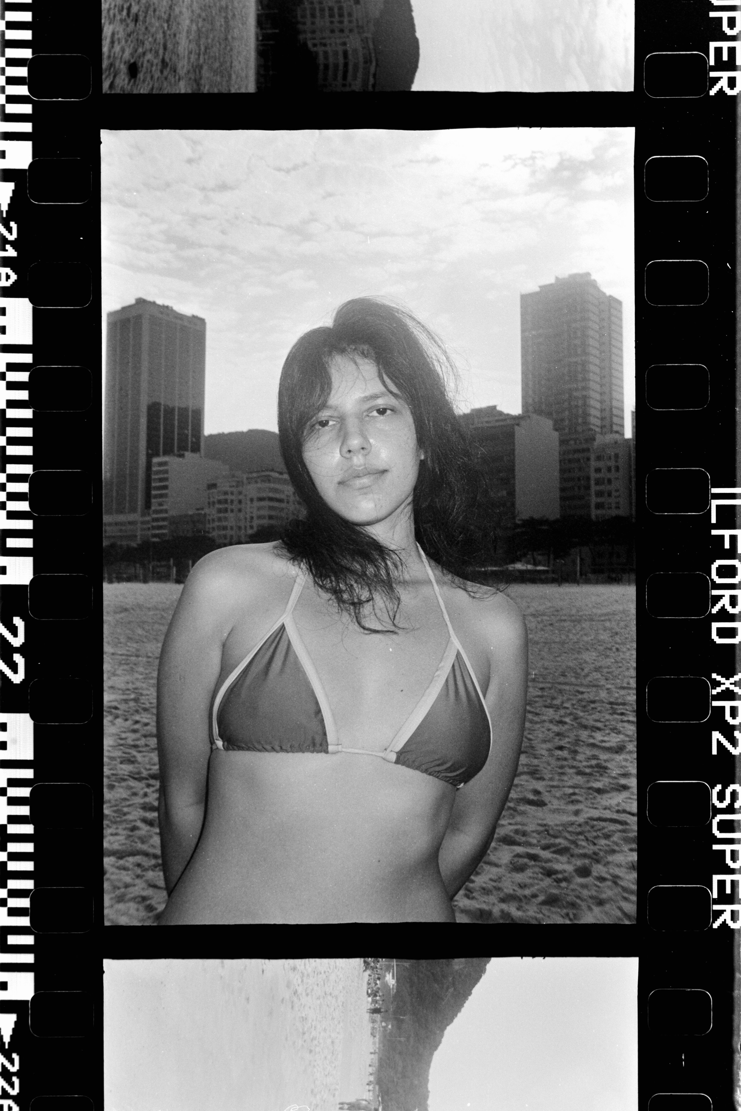 Black and white photo of a woman in a bikini standing on a beach with city buildings in the background.
