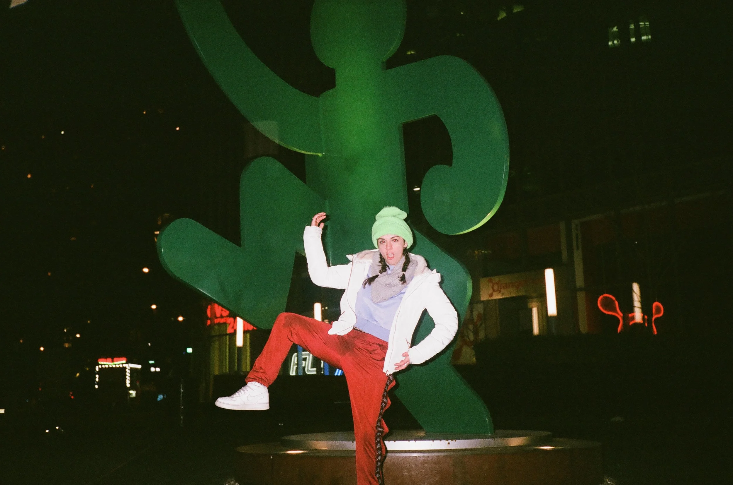 A young woman with a green beanie, white jacket, grey hoodie, and red pants posing in front of a large green dollar sign sculpture at night.