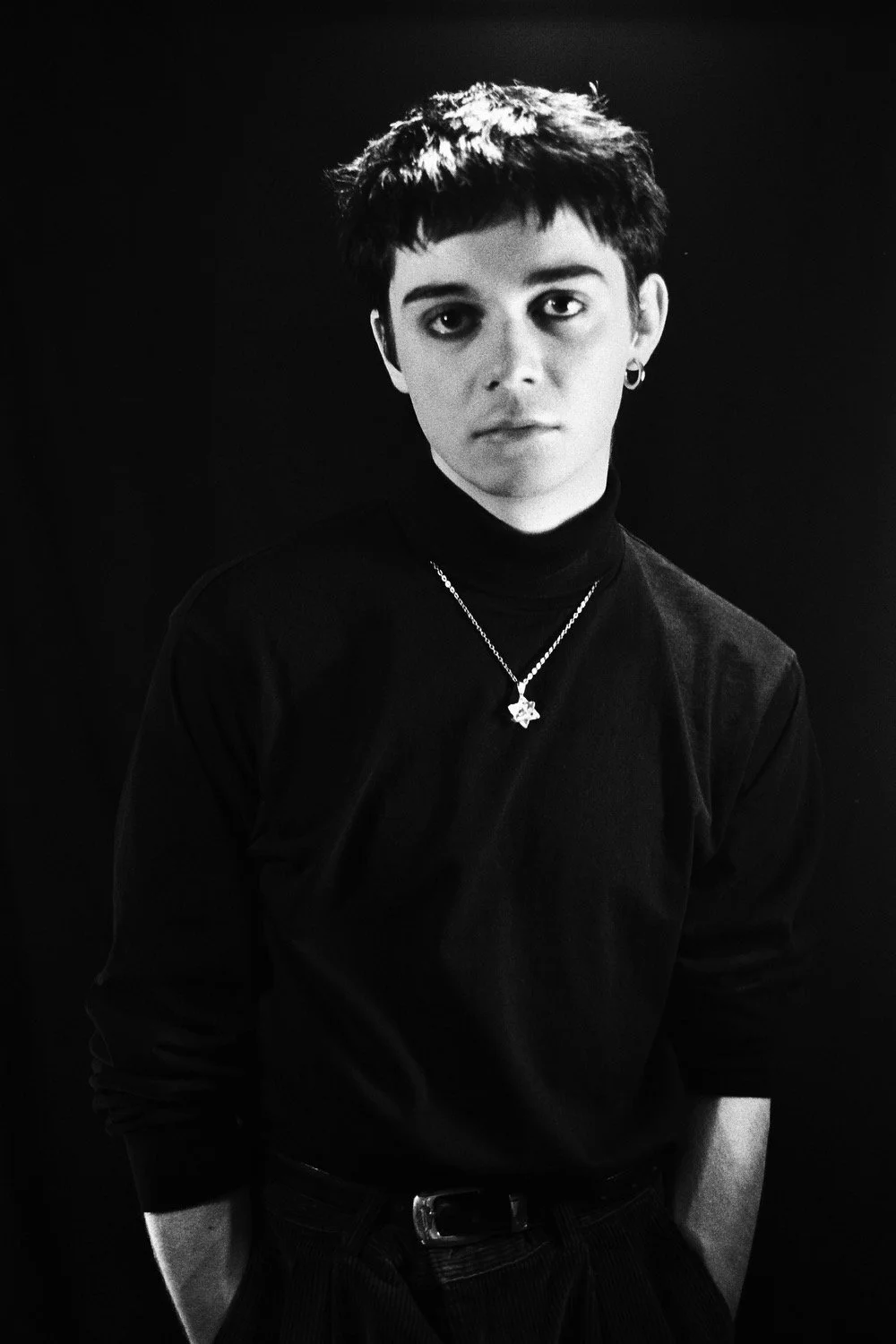 Black and white portrait of a young person with short, tousled hair, wearing a black turtleneck, hoop earrings, a necklace with a star-shaped pendant, and dark pants, standing against a dark background.