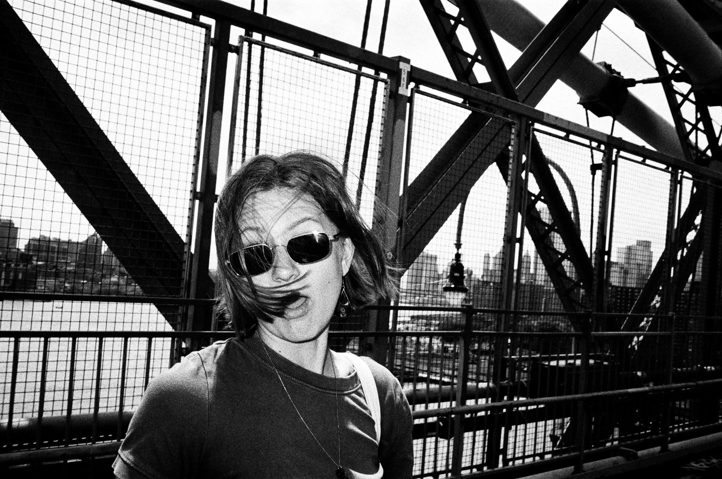 A woman with short hair wearing sunglasses and a t-shirt is posing with her mouth open in front of a metal bridge structure, cityscape, and water in a black-and-white photo.