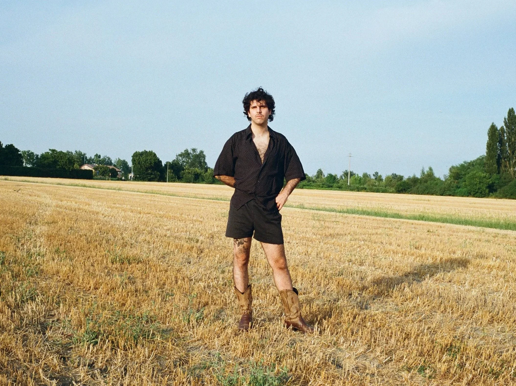 A young man with curly dark hair stands in a harvested field, wearing a black shirt, black shorts, and cowboy boots, with a backdrop of trees, houses, and a clear sky.