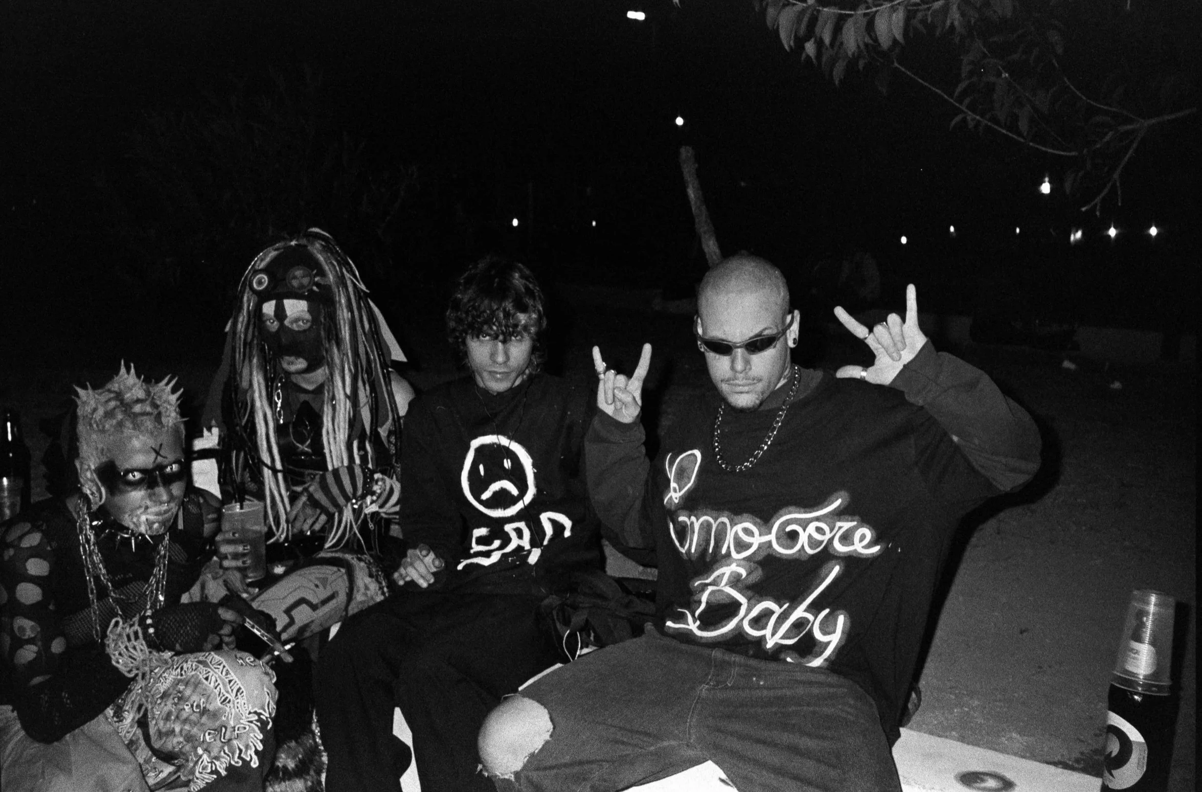 Group of four young adults at night, dressed in punk and goth fashion, sitting outdoors. They are making hand gestures, and one is holding a drink. The photo is black and white.
