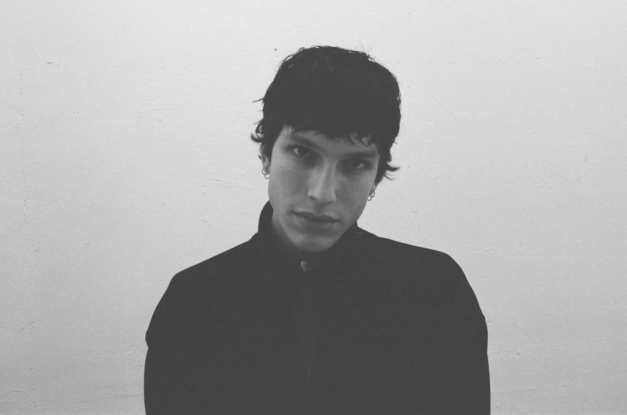 Black and white portrait of a young person with short, dark, wavy hair, wearing a zip-up jacket, looking directly at the camera with a serious expression against a plain wall background.