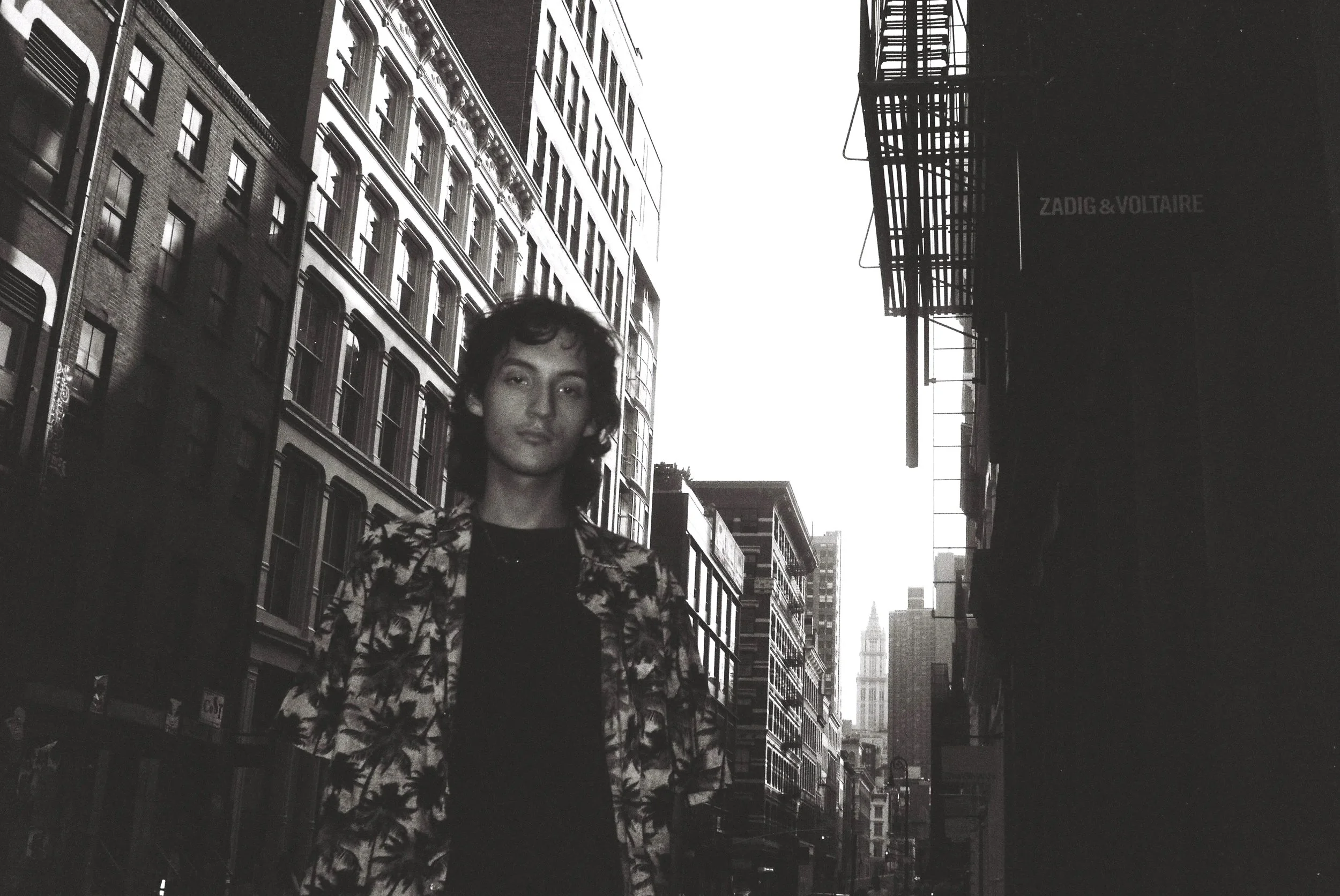 Black and white photo of a young woman standing on a city street with tall buildings around her, time appears to be either early morning or late evening.