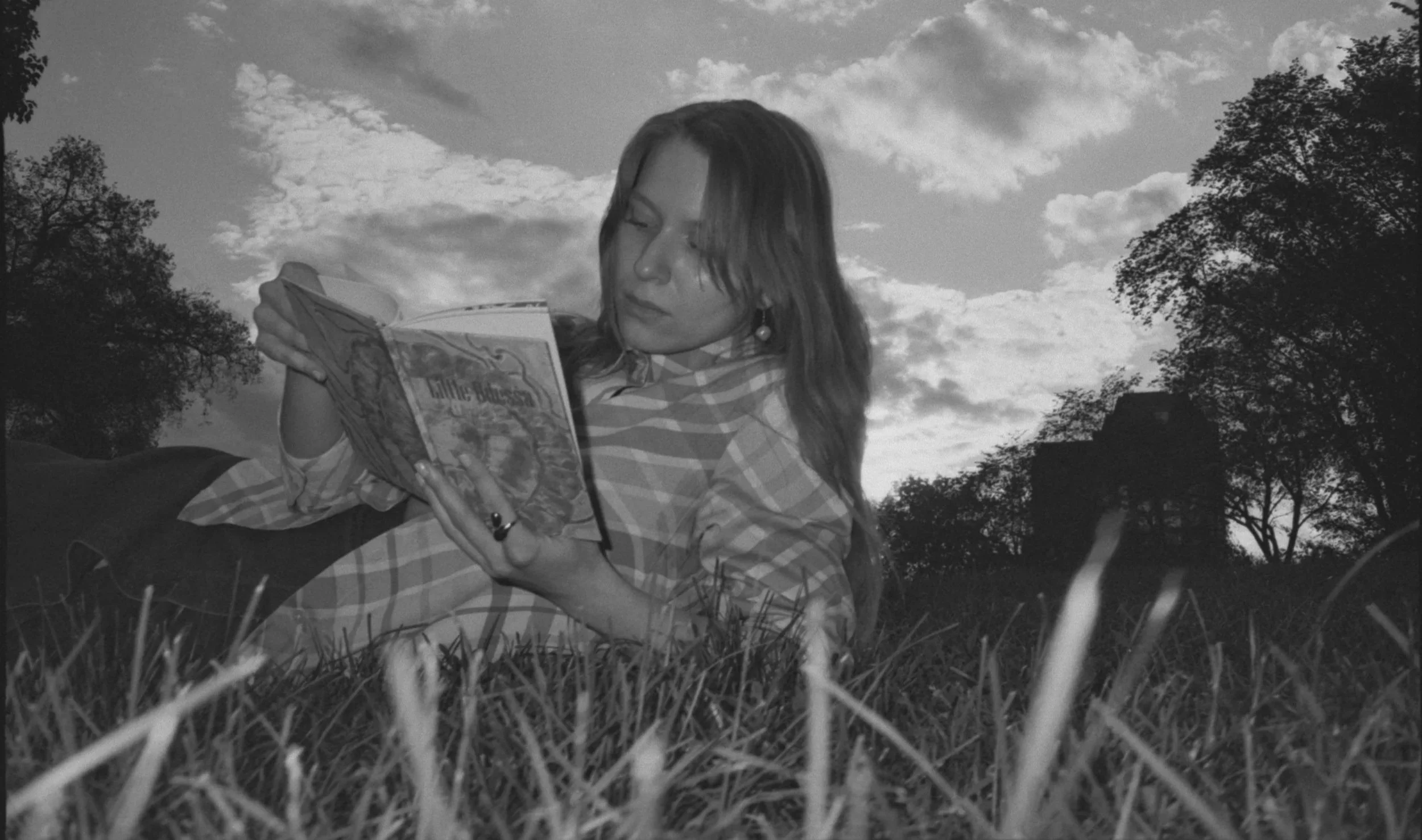 A woman lying on the grass outdoors, reading a book with a cloudy sky and trees in the background.