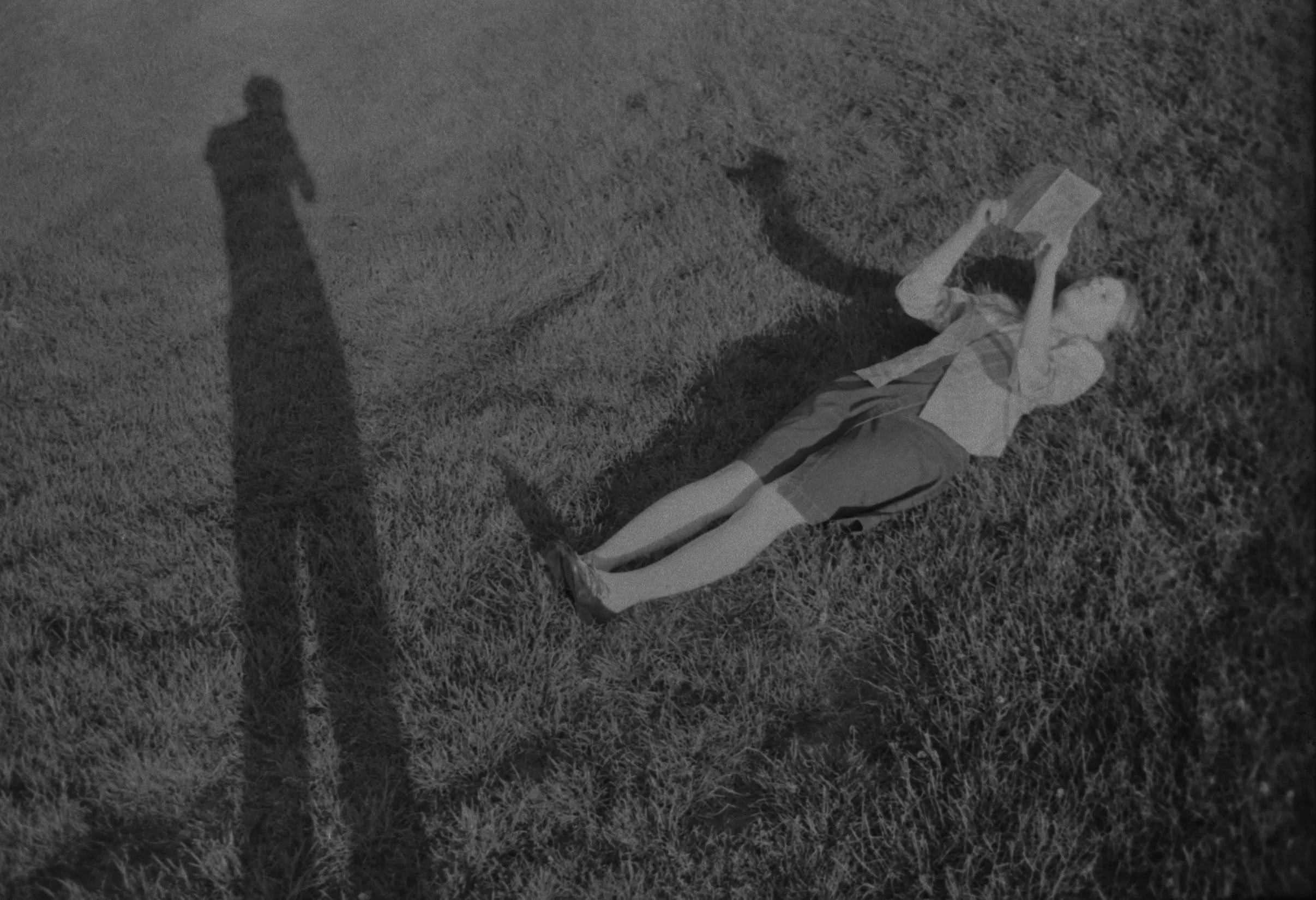 Child lying on grass, reading a book at night, cast shadow of an adult figure nearby.