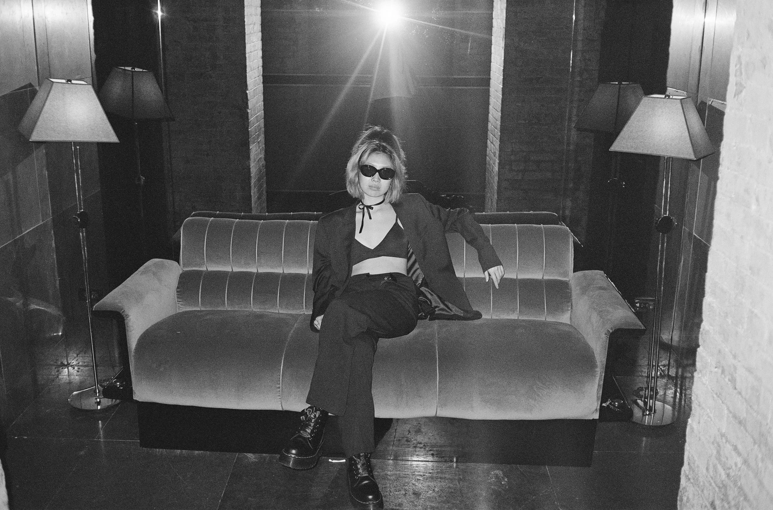 A woman sitting on a sofa in a dimly lit room with two lamps on either side. She is wearing sunglasses, a black top, a blazer, and boots, with one leg crossed over the other.