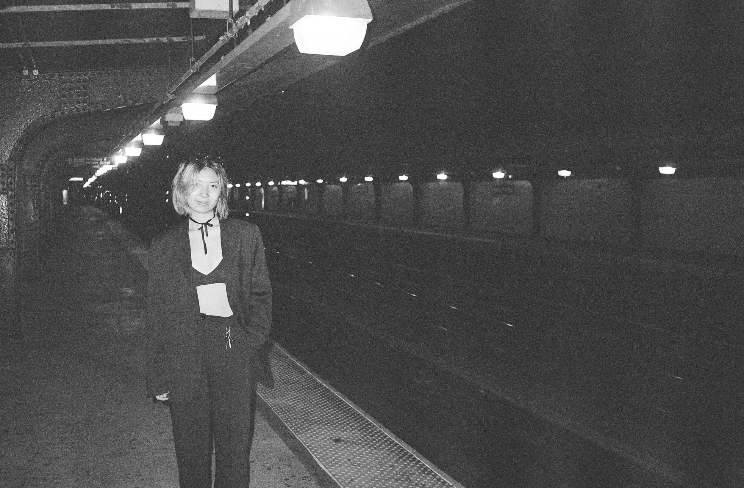 A woman with blonde hair, wearing a black blazer and pants, stands on a subway platform at night. The subway tracks and dark tunnel are visible behind her.