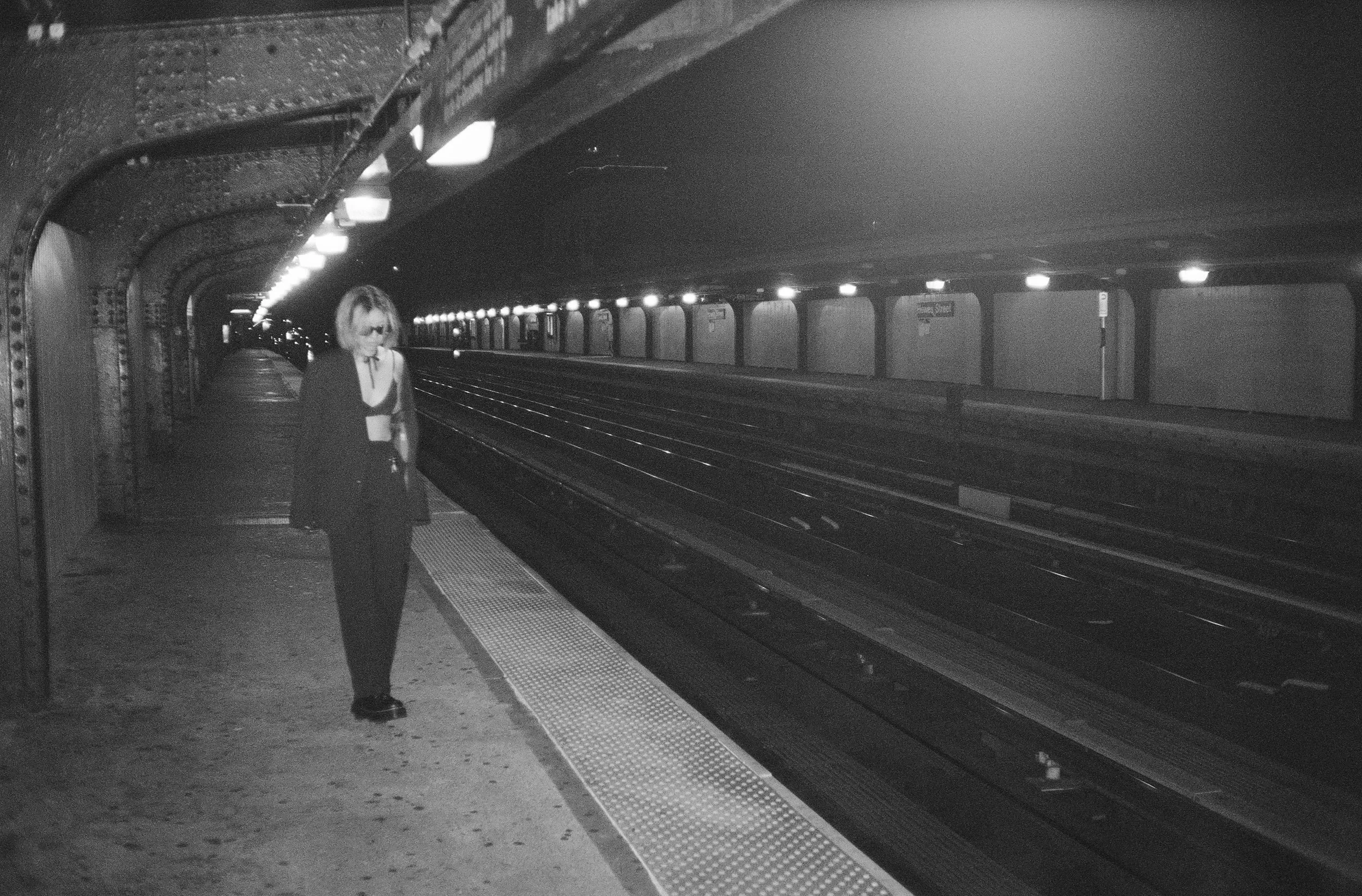 A woman standing on a subway platform next to empty tracks in an underground station at night, with artificial lighting along the ceiling.