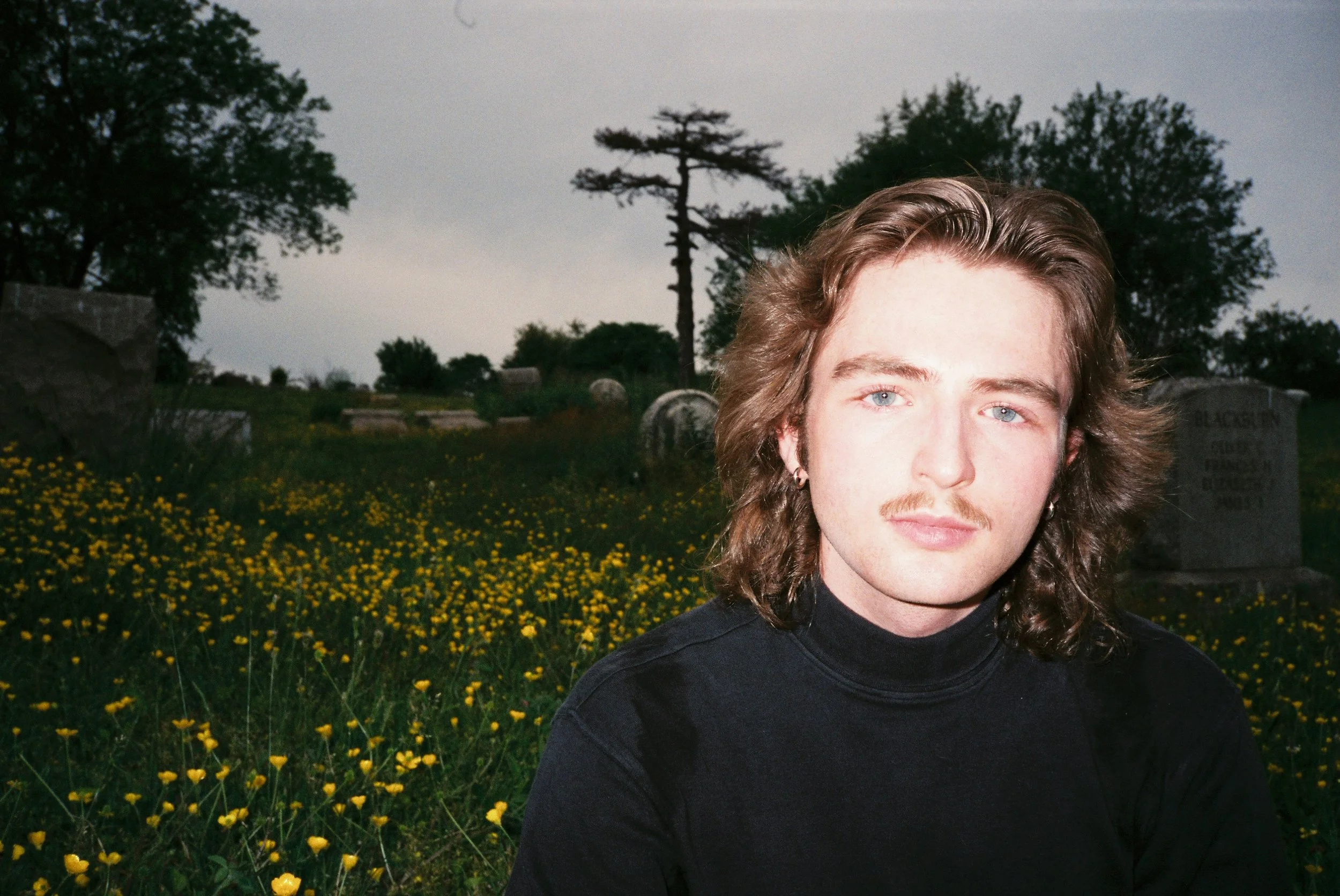 A young man with shoulder-length brown hair, blue eyes, and a mustache, wearing a black turtleneck shirt, sitting in a field of yellow flowers with trees and gravestones in the background.