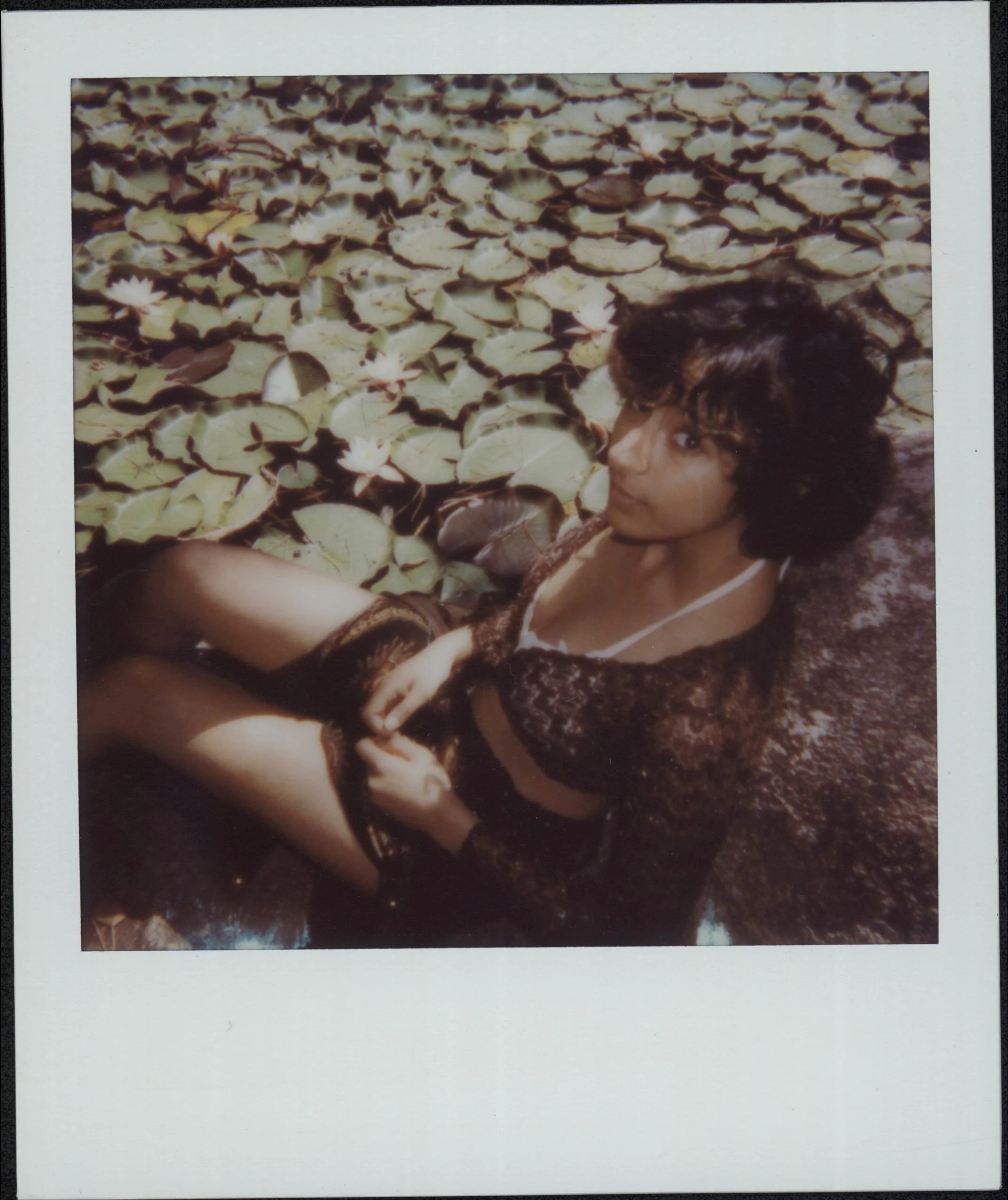 Girl with dark curly hair reclining on a large rock among lily pads and white water lilies in a pond.