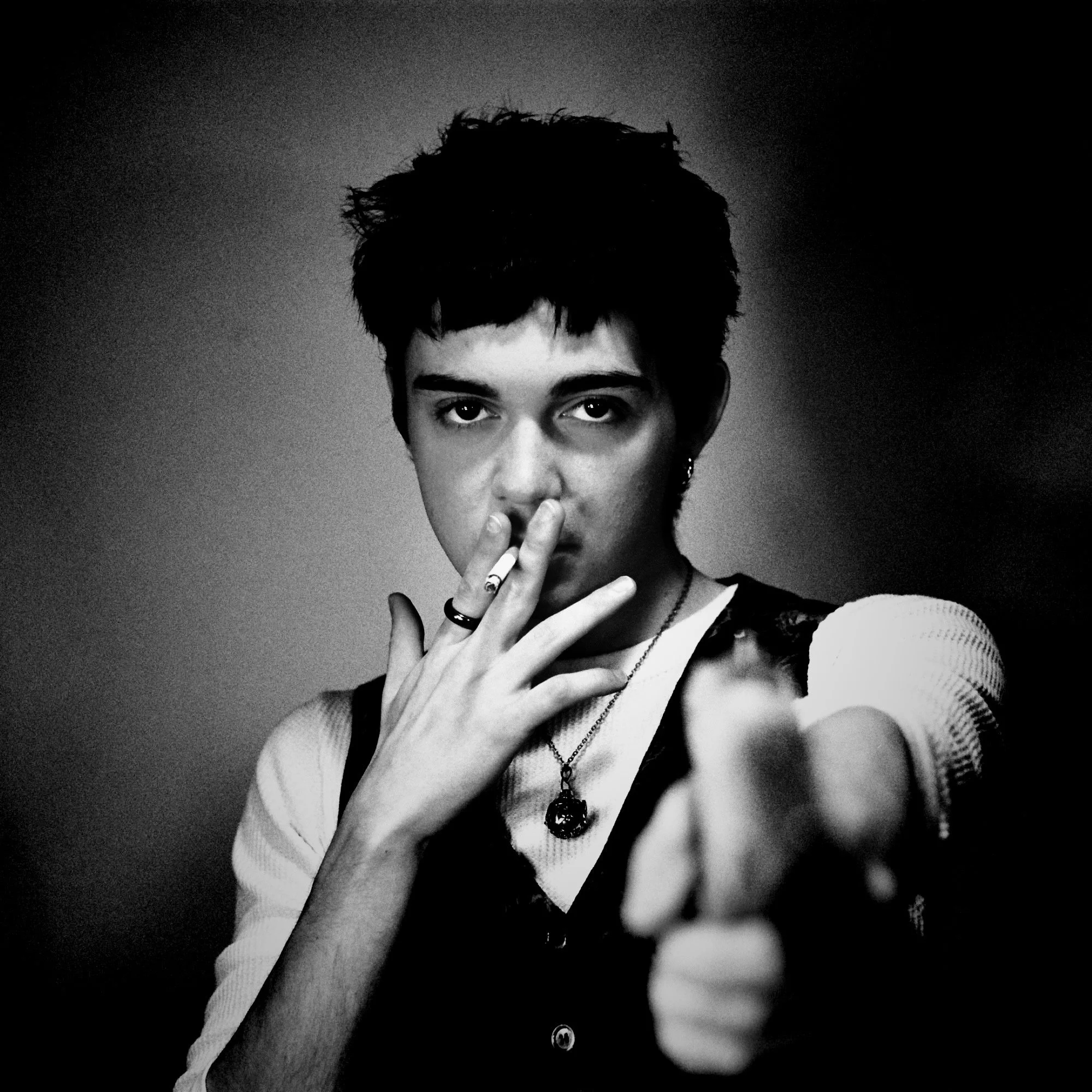 Black and white portrait of a young man with dark, messy hair, holding a cigarette near his lips, wearing rings and necklaces, with a serious expression, extending his hand toward the camera.