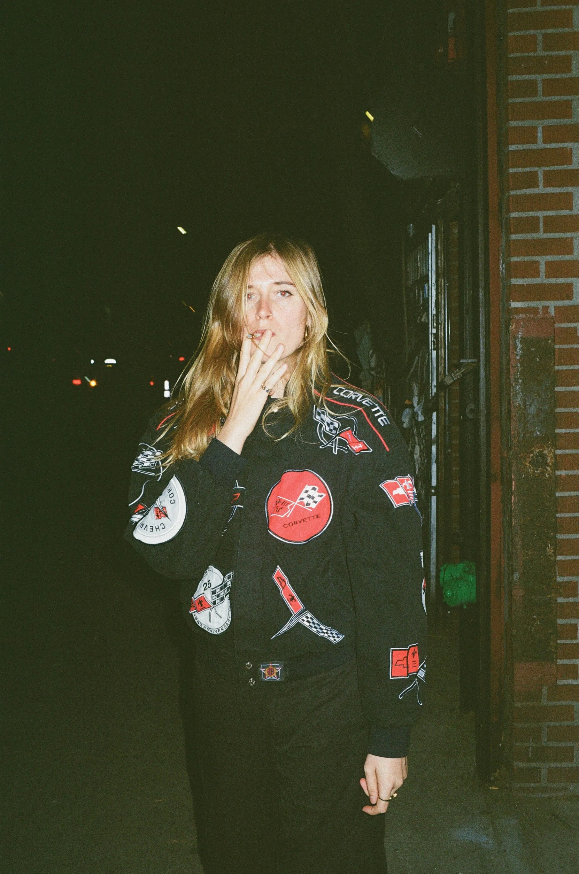 A woman with blonde hair standing outdoors at night, smoking a cigarette, wearing a black jacket with various Corvette logos and patches, standing next to a brick wall.