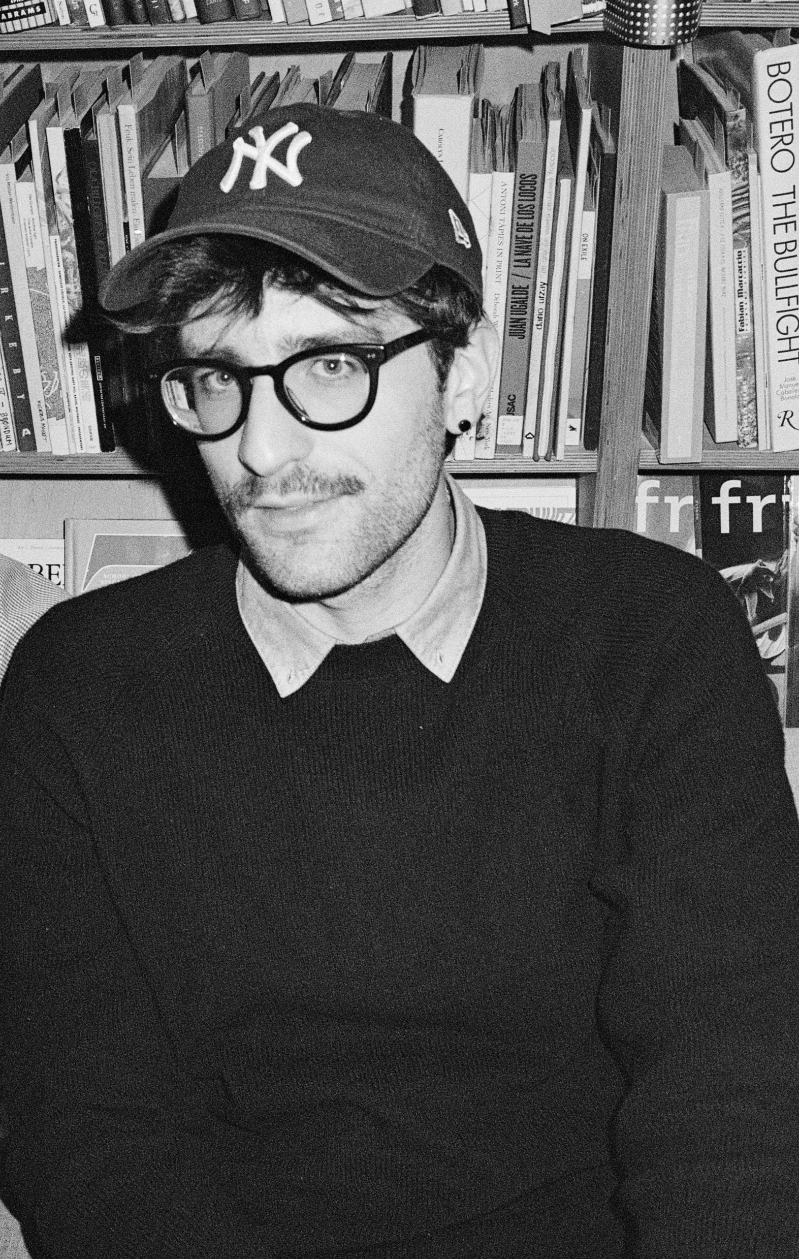A man with glasses, a mustache, and a beard wearing a New York Yankees cap and a collared shirt under a sweater, sitting in front of a bookshelf filled with various books.
