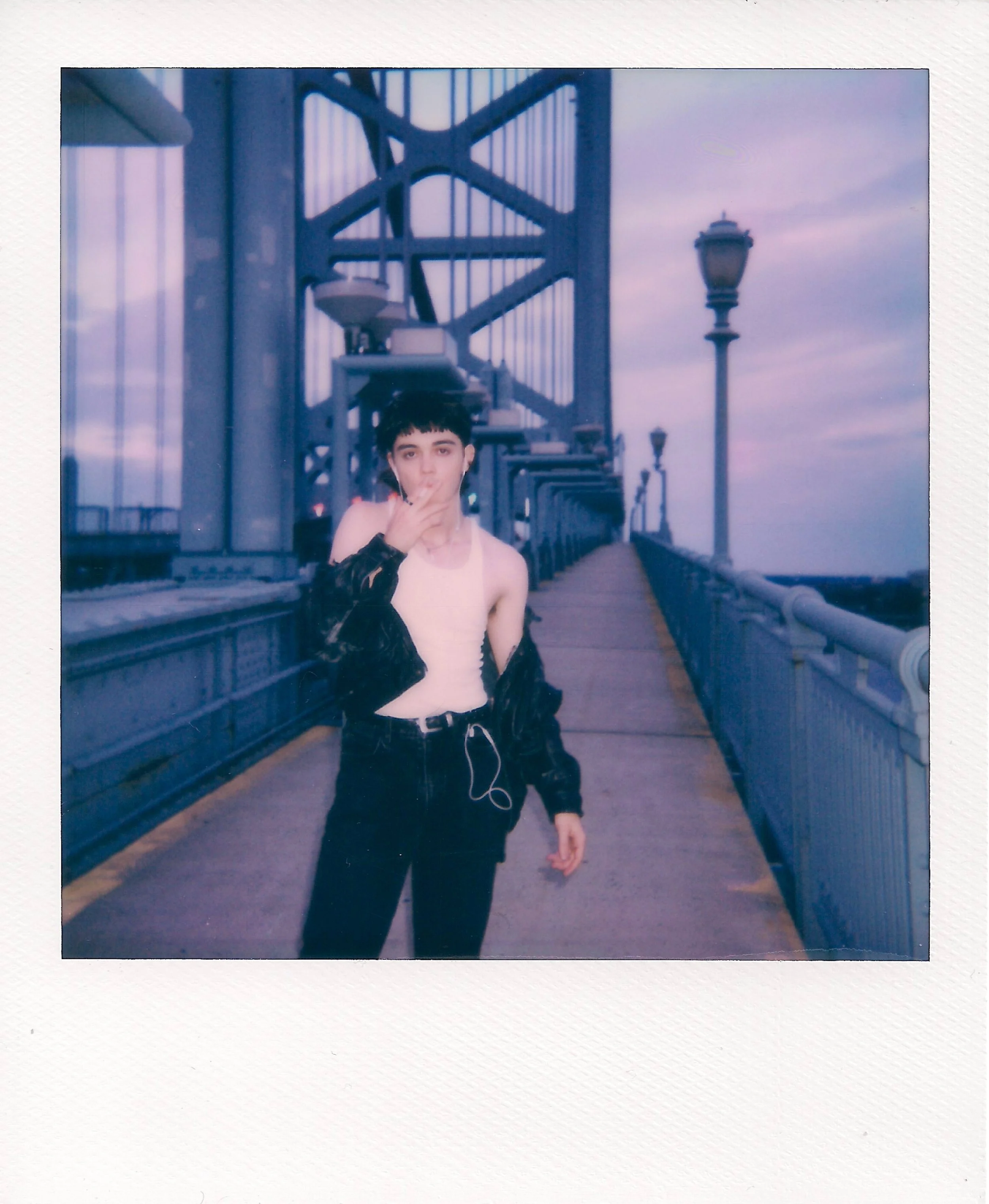 A young person with short dark hair standing on a walkway of the Brooklyn Bridge with the city skyline in the background during dusk.
