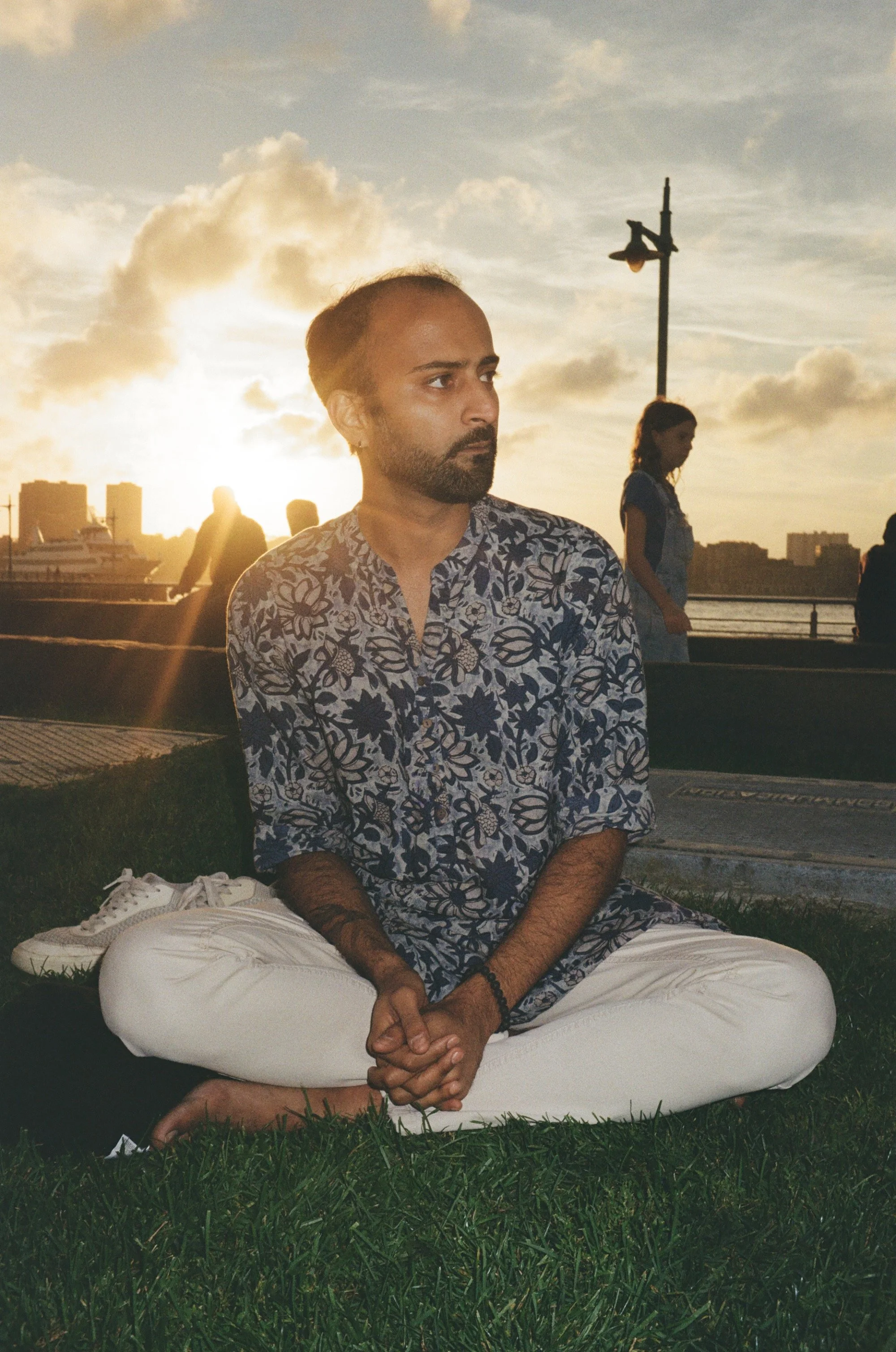 A man sitting cross-legged on the grass near a waterfront during sunset, with two women and city skyline in the background.