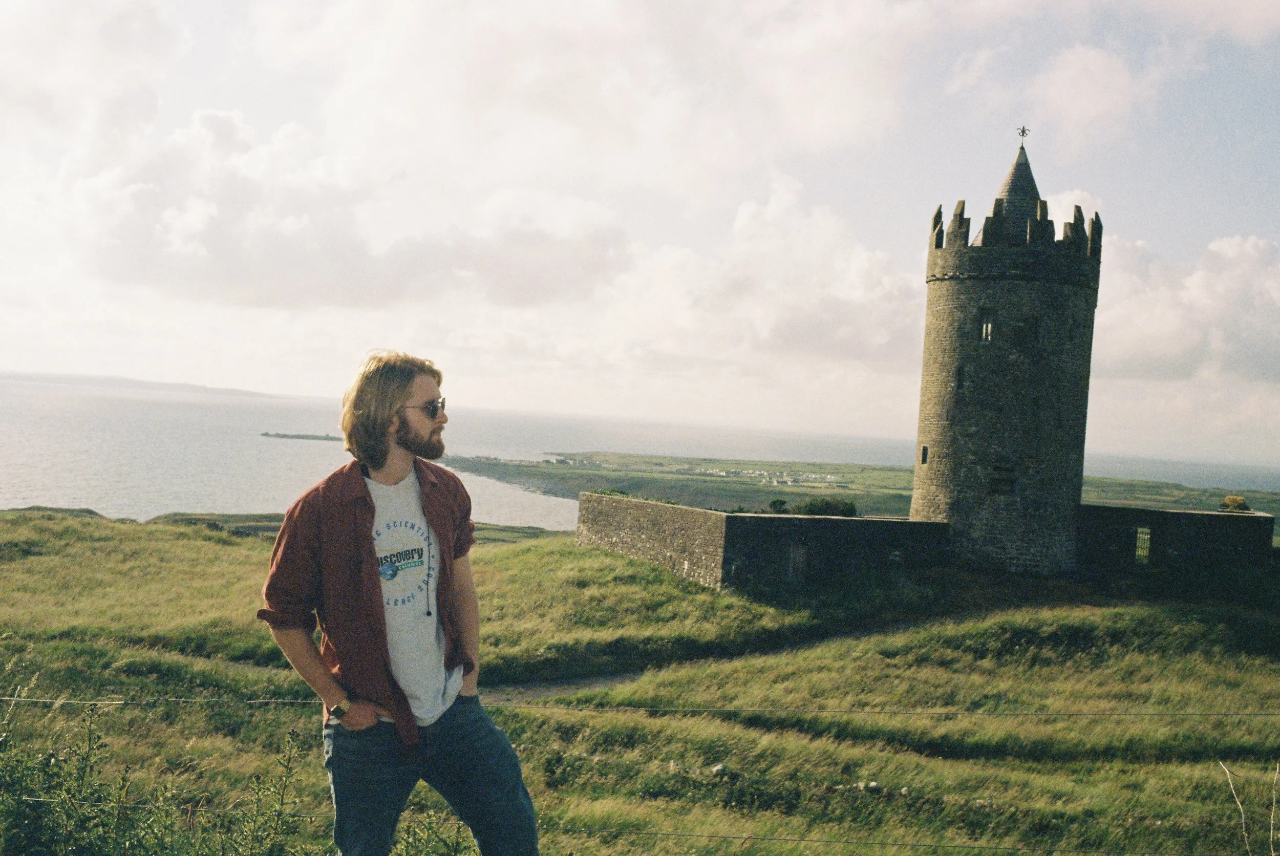 A man with long hair, beard, sunglasses wearing a maroon jacket and white t-shirt standing on green grass near a historic stone tower with a body of water and a cloudy sky in the background.