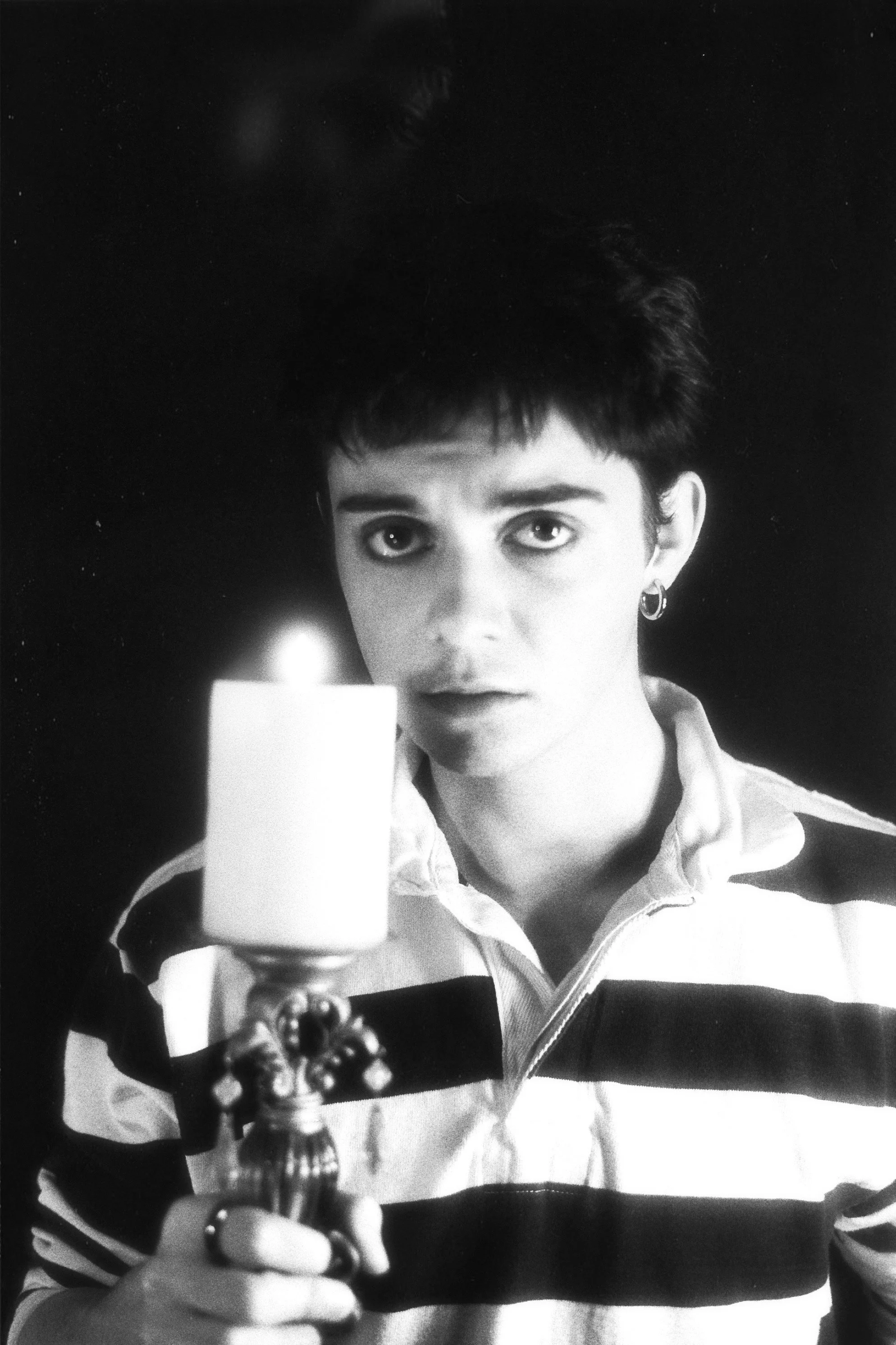 A young woman with short dark hair and earrings, holding a lit candle in a candle holder, looking directly at the camera with a serious expression.