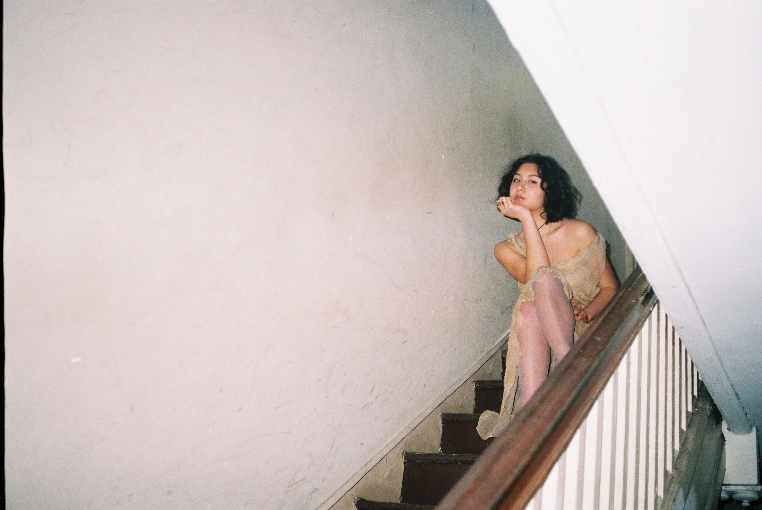 A woman with curly black hair sitting on a staircase, resting her chin on her hand, wearing a sheer beige dress and pink fishnet stockings.