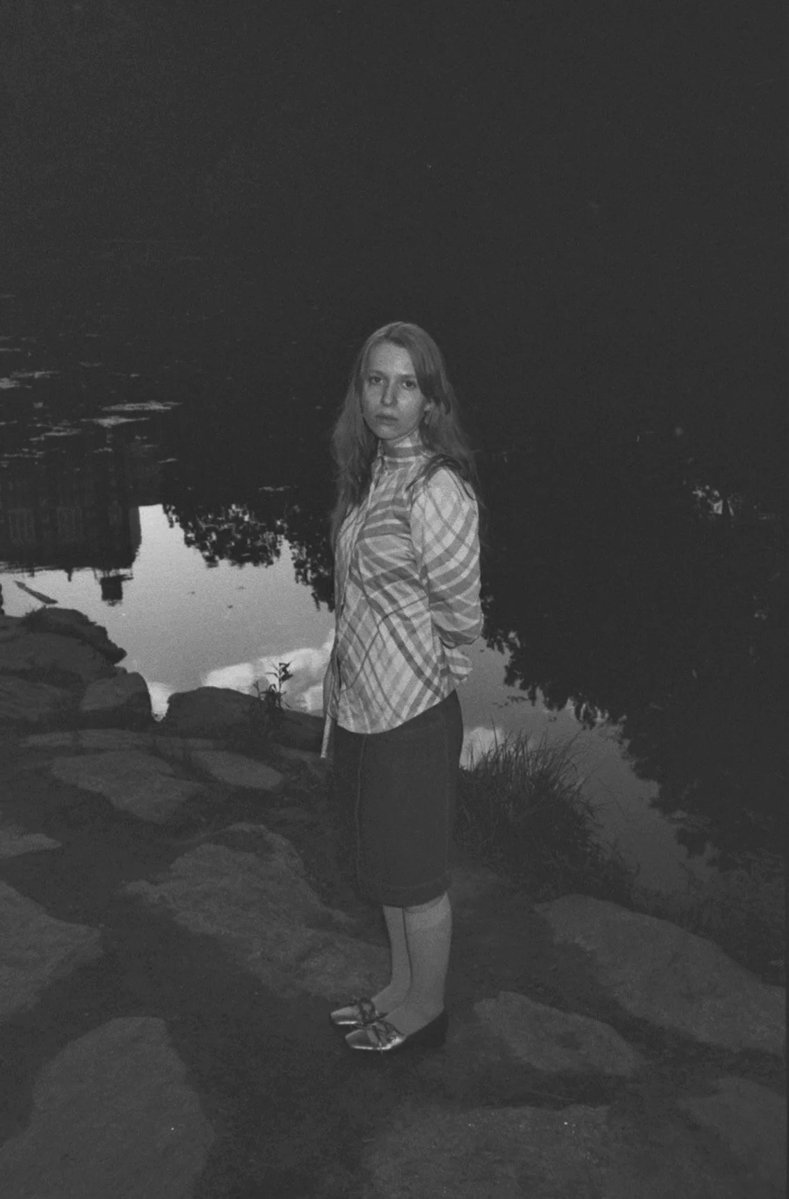 A woman standing by a body of water at night, wearing a checkered shirt, skirt, and flats, with trees and a building reflected in the water.